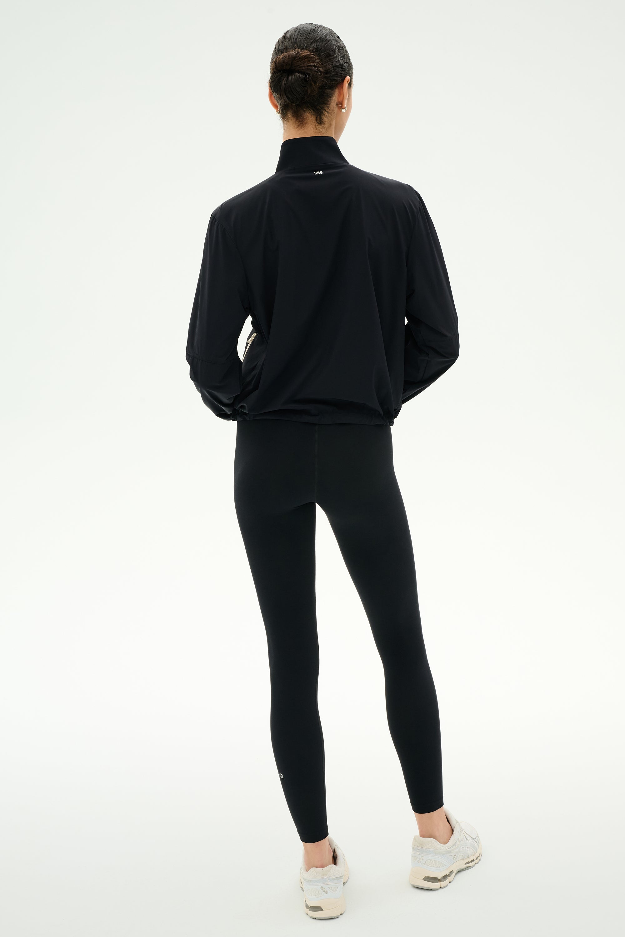 A person stands with their back to the camera, wearing the SPLITS59 Lyle Woven Nylon Pullover in Black/Creme, paired with black spandex leggings and white sneakers against a plain light background.