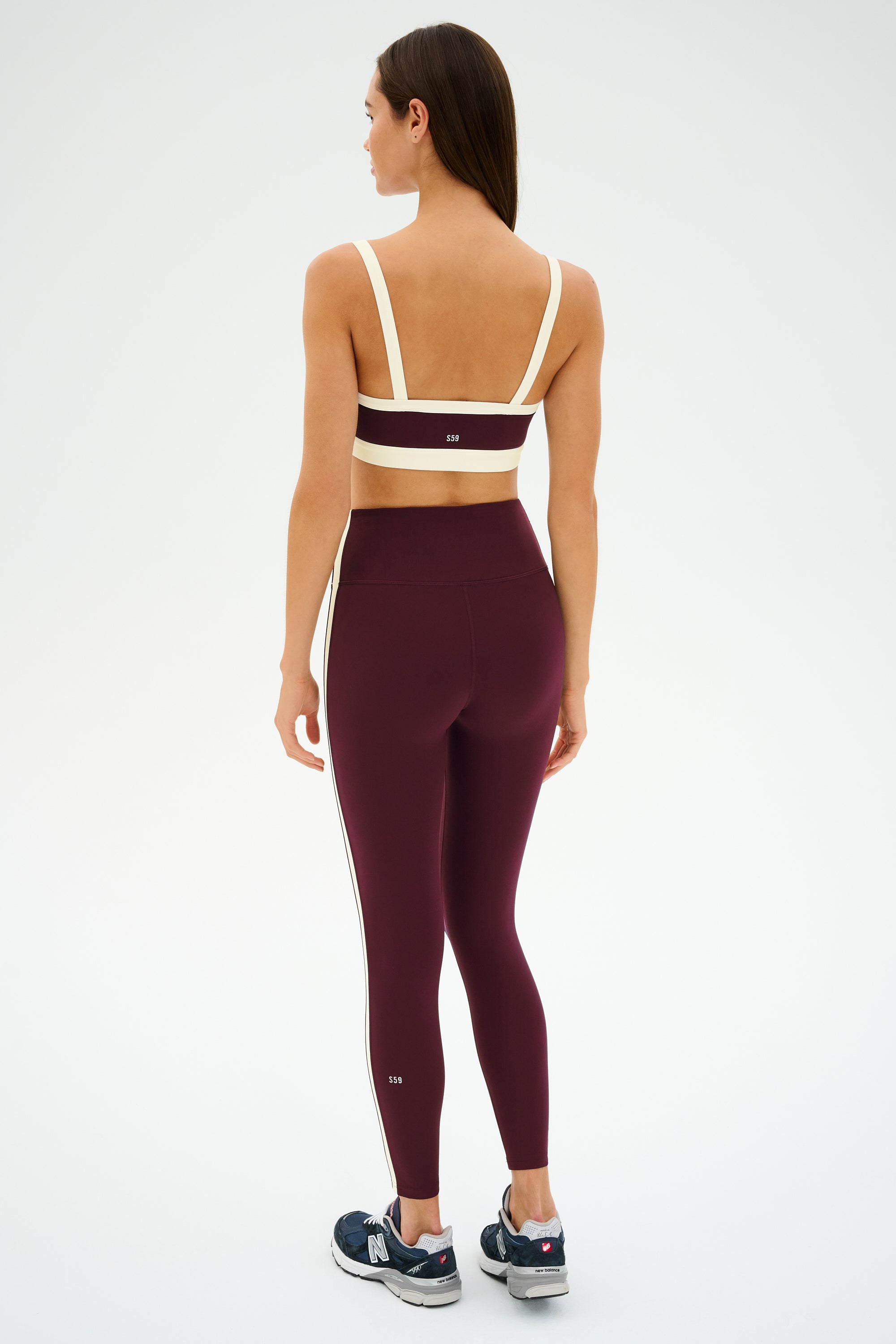 A woman faces away, wearing the SPLITS59 Monah Rigor Bra in Dark Cherry/Creme, paired with high-waisted maroon spandex leggings with a side stripe and navy sneakers against a plain background.