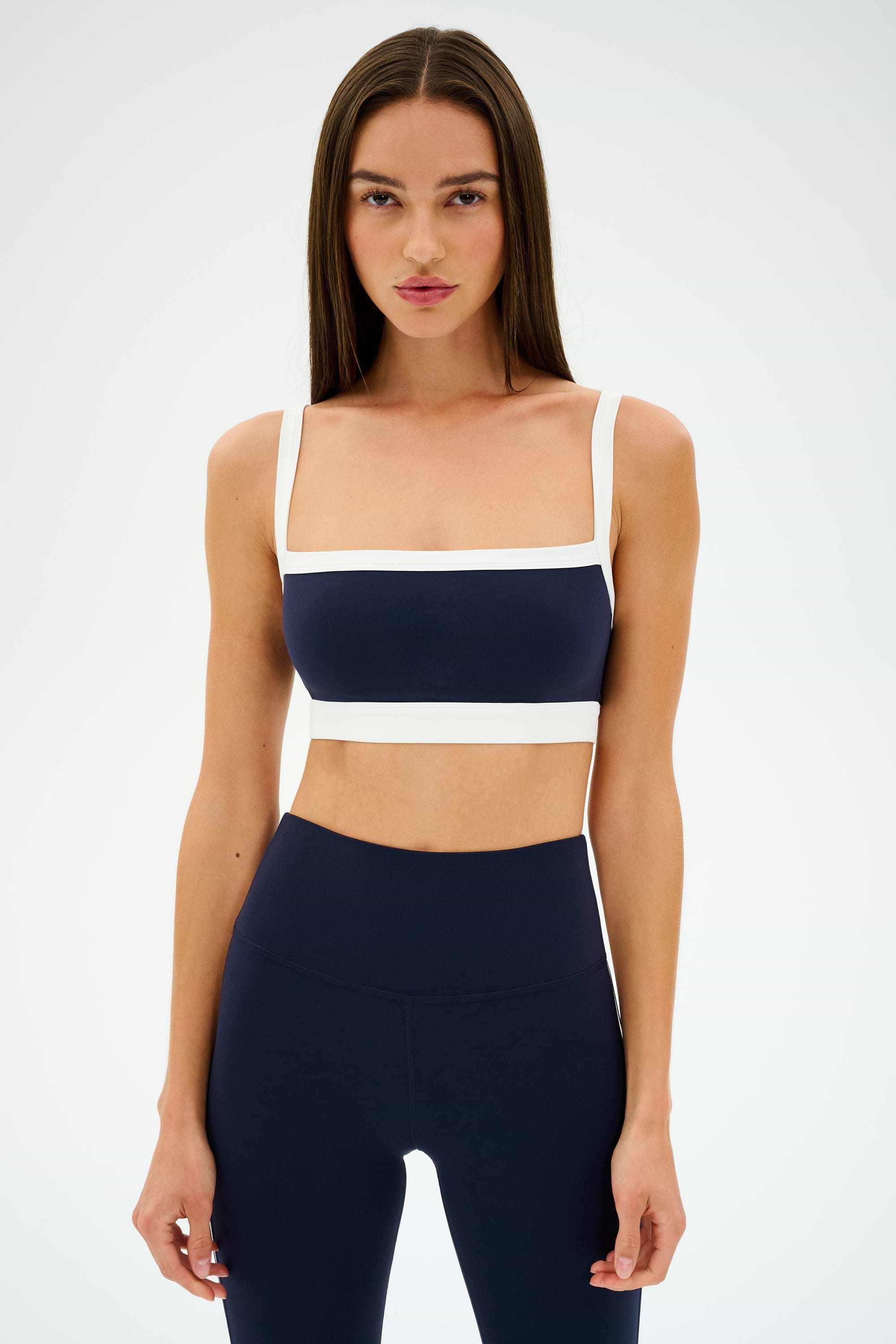 Against a plain background, a person in an indigo and white athletic outfit exudes a retro sport vibe, featuring the SPLITS59 Monah Rigor Bra for solid support.