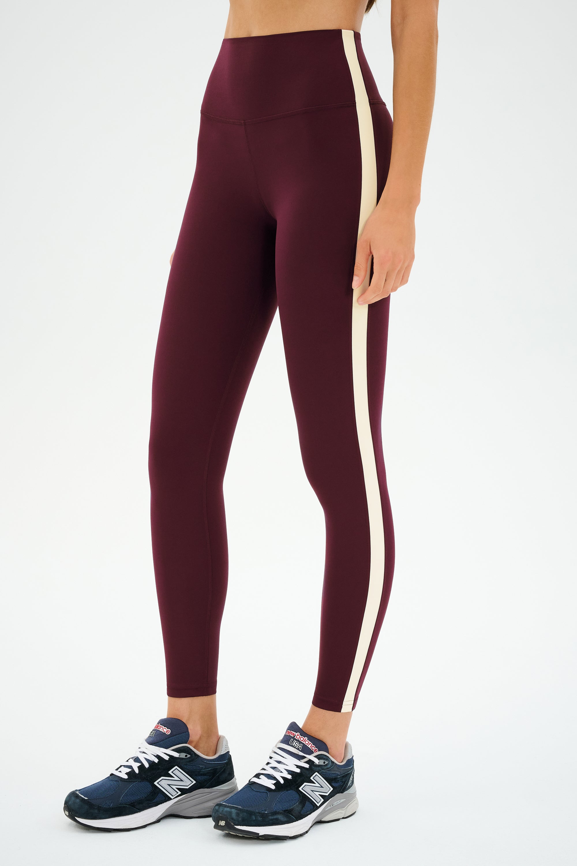 A person stands against a white background wearing SPLITS59 Clare High Waist Rigor 7/8 leggings in Dark Cherry/Creme with maroon Rigor fabric and beige side stripe, paired with dark blue New Balance sneakers.