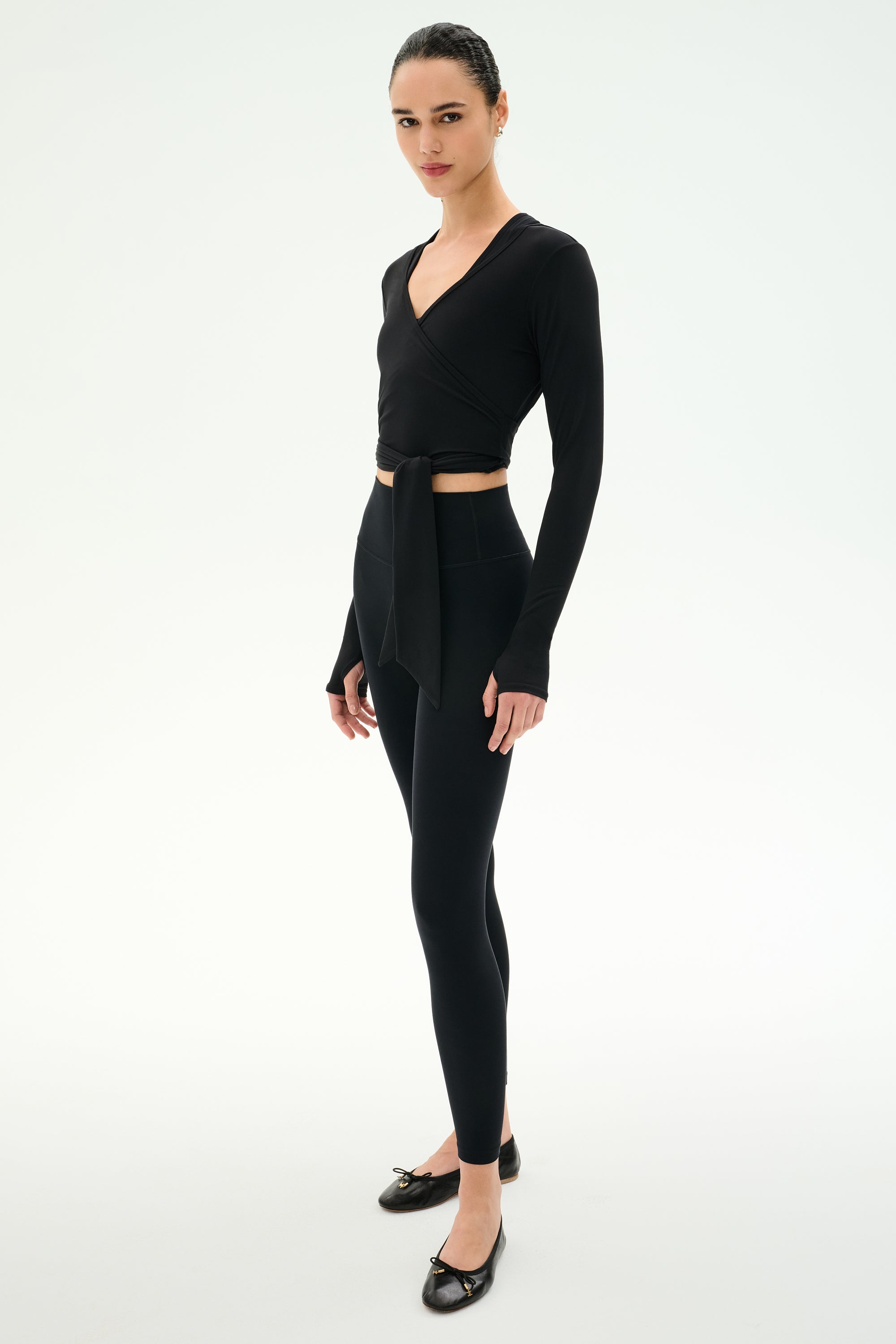 A person stands against a white background wearing the SPLITS59 Bella Airweight Lite Wrap in black, paired with black spandex leggings and black ballet flats.