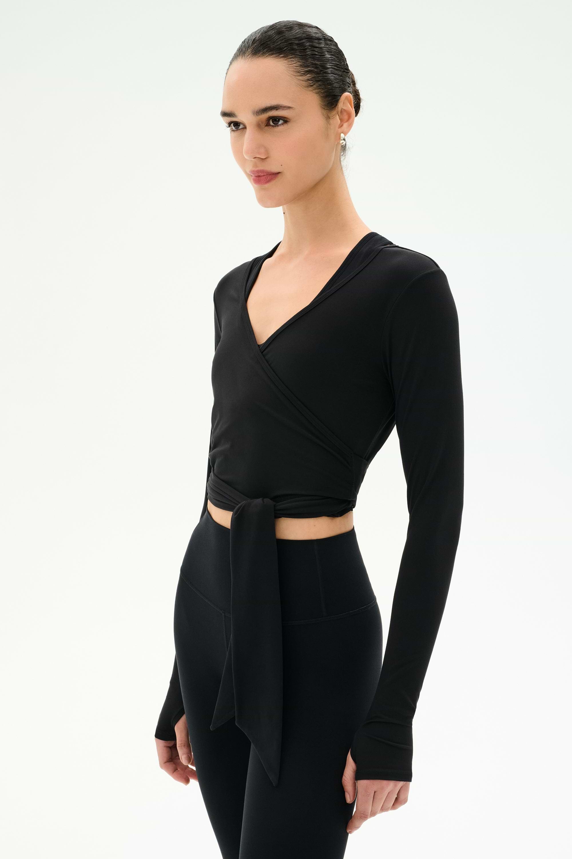 A woman stands against a plain white background wearing the SPLITS59 Bella Airweight Lite Wrap in black, a stylish long-sleeve spandex top with a front tie, paired with black high-waisted leggings.