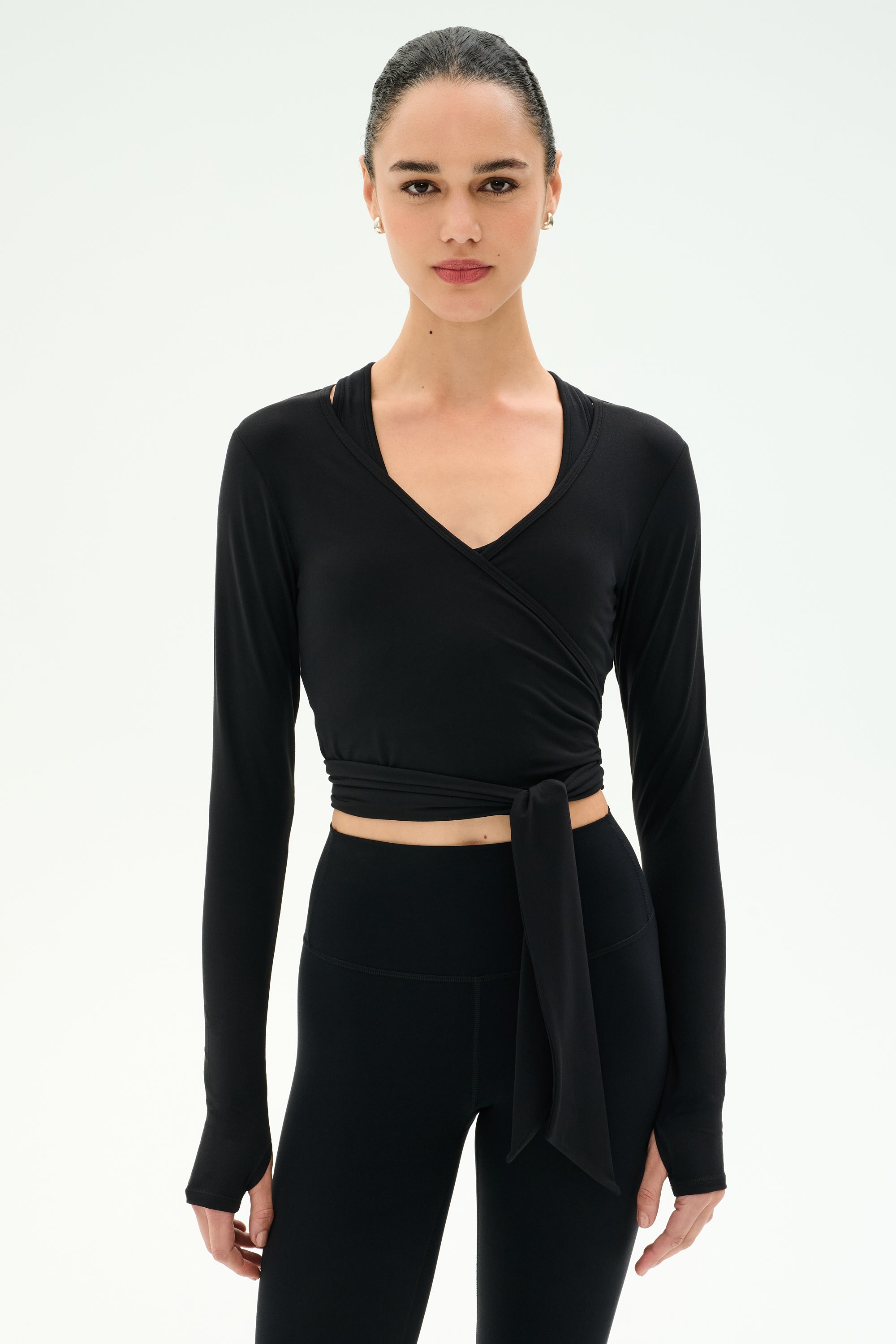 A woman wears the SPLITS59 Bella Airweight Lite Wrap in black—a long-sleeve, wrap-style crop top—paired with high-waisted black leggings, posed against a plain white background.