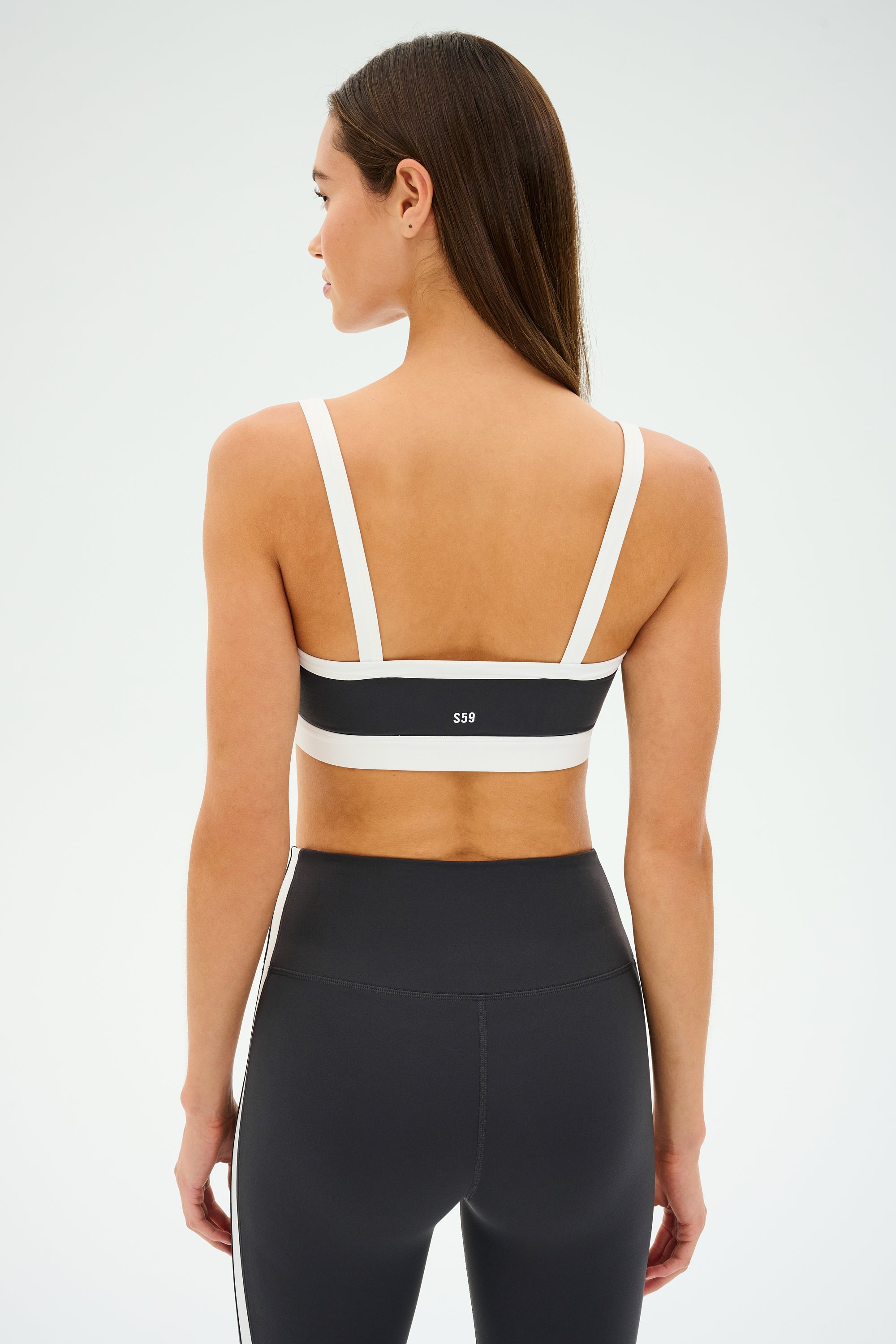 A person confidently stands with their back to the camera, wearing a SPLITS59 Monah Rigor Bra in Graphite/White and matching leggings, ready for gym workouts against a light background.