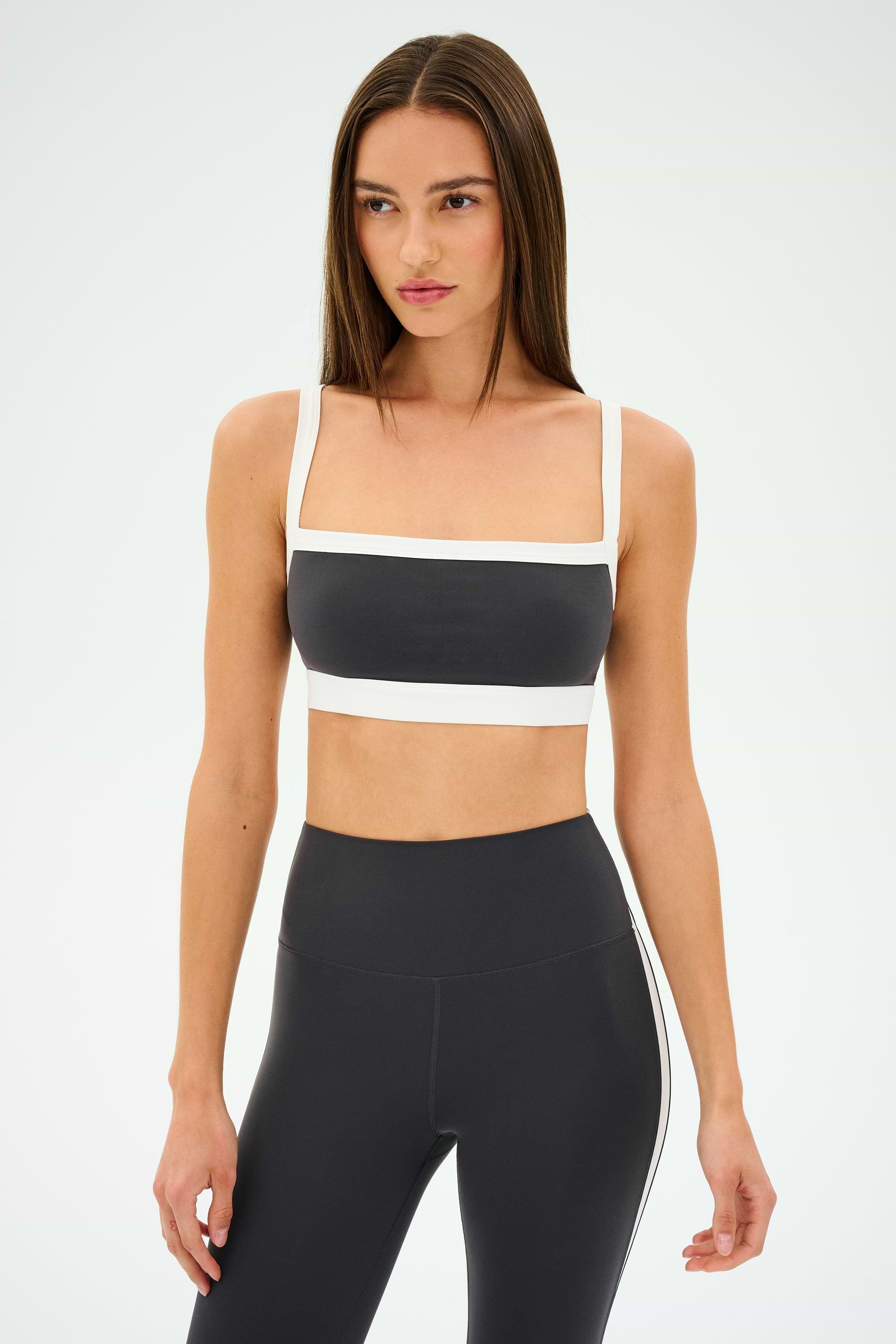 Poised against a plain backdrop, a person wearing a chic SPLITS59 Monah Rigor Bra in Graphite/White and black leggings is ready to conquer their gym workouts.