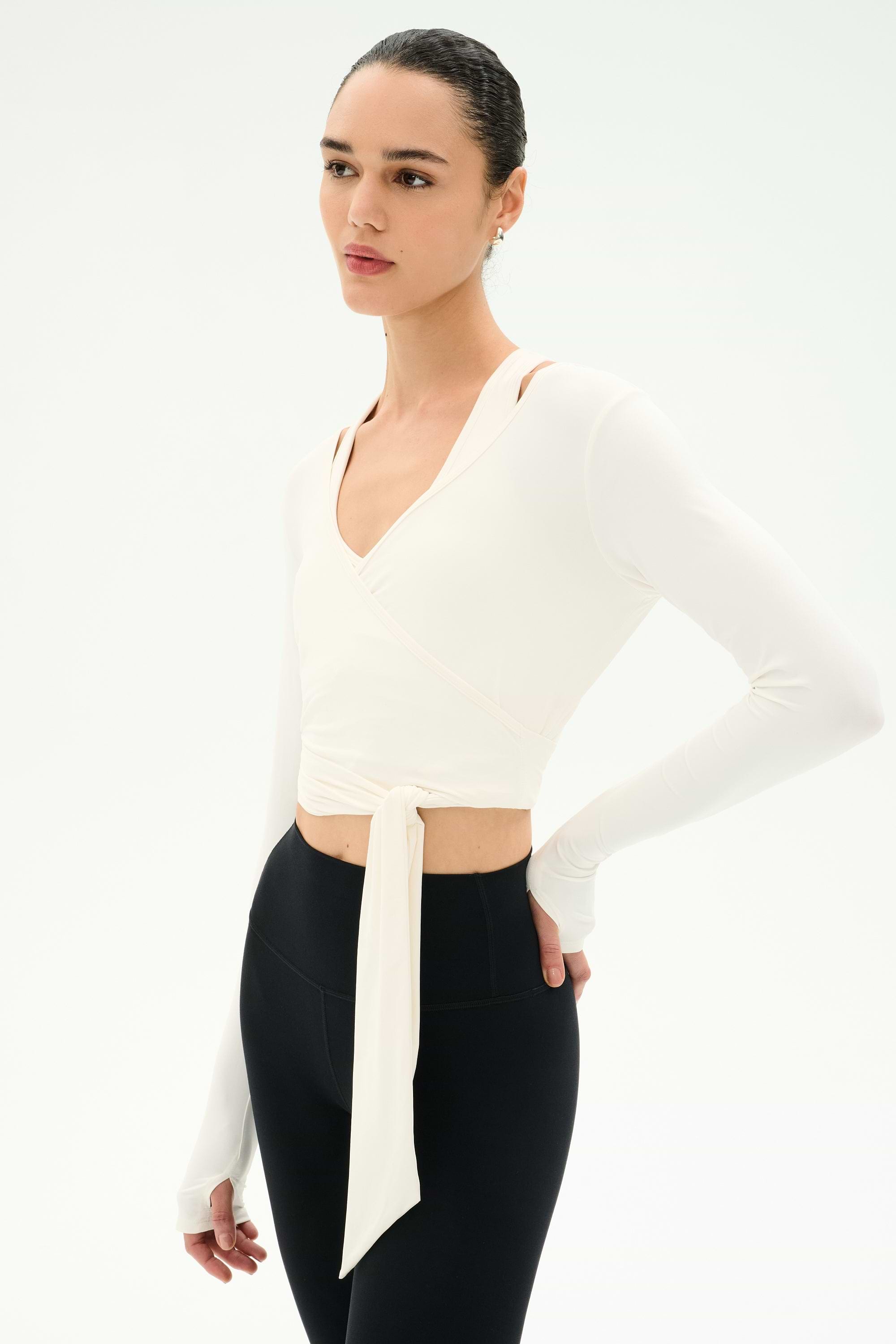 A person wearing the SPLITS59 Bella Airweight Lite Wrap in white stands against a plain white background, paired with black high-waisted nylon-spandex leggings.