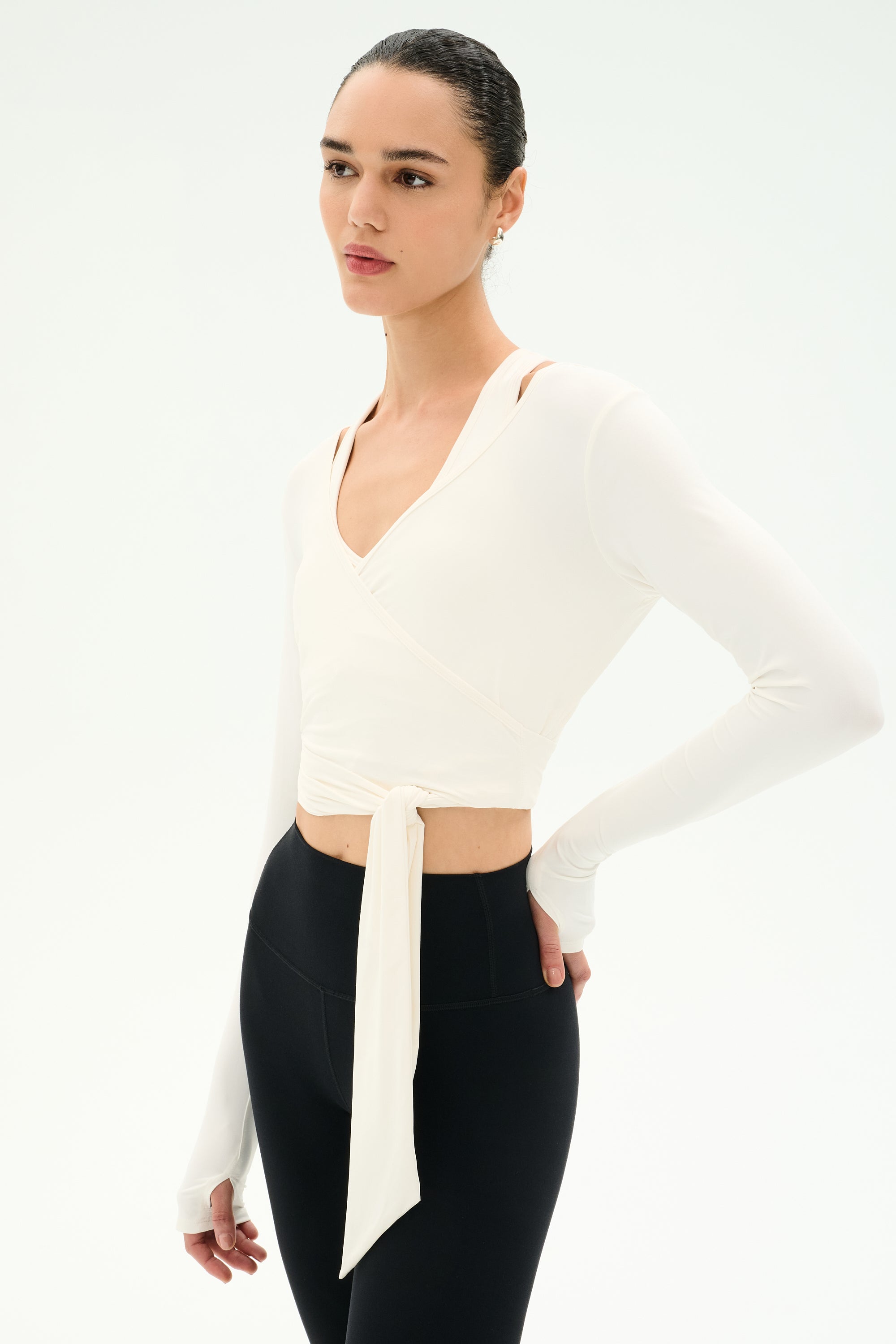 A person wearing the SPLITS59 Bella Airweight Lite Wrap in white stands against a plain white background, paired with black high-waisted nylon-spandex leggings.