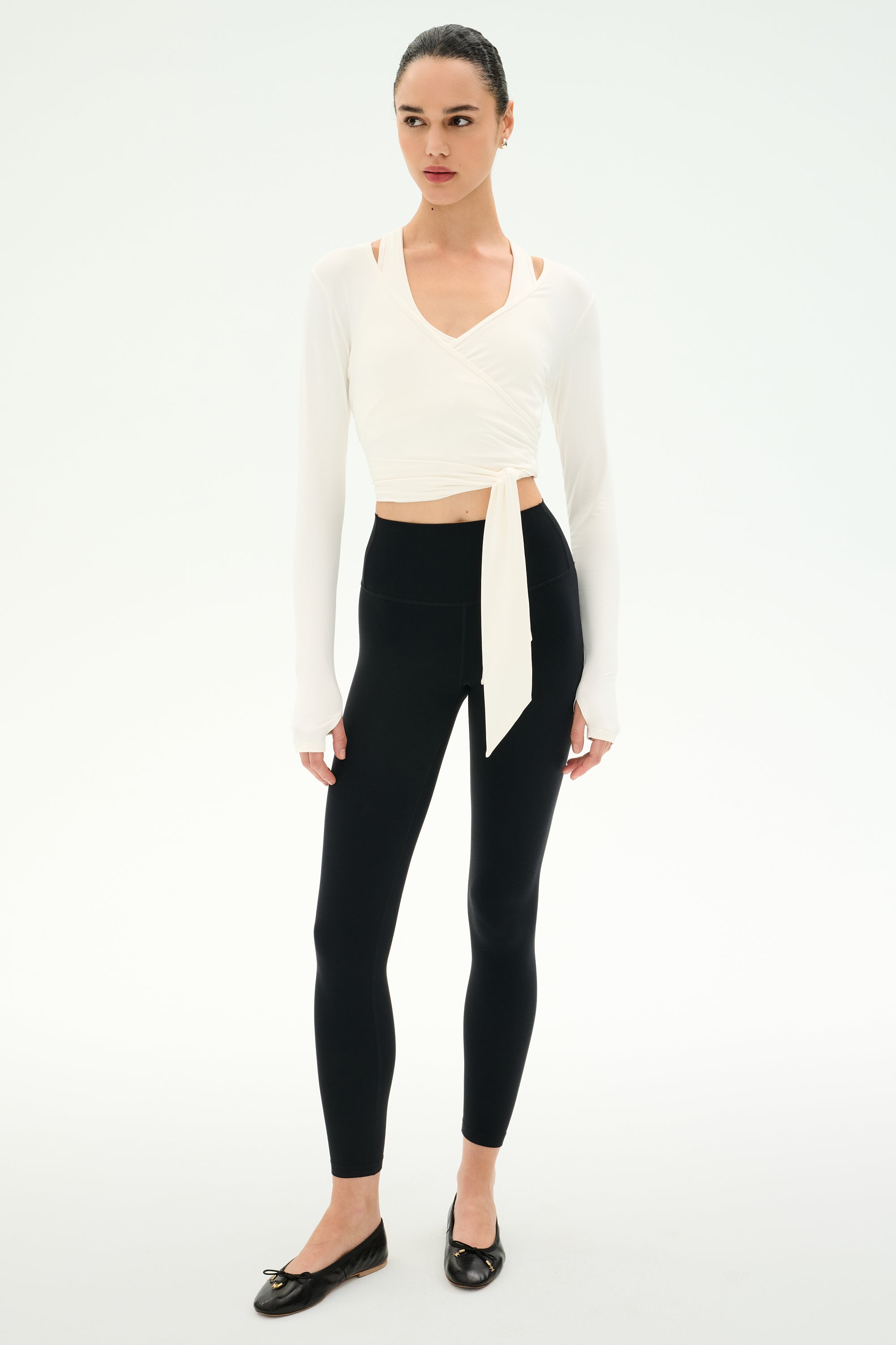 A woman wears the SPLITS59 Bella Airweight Lite Wrap in white, paired with black leggings and ballet flats, standing against a plain white background.