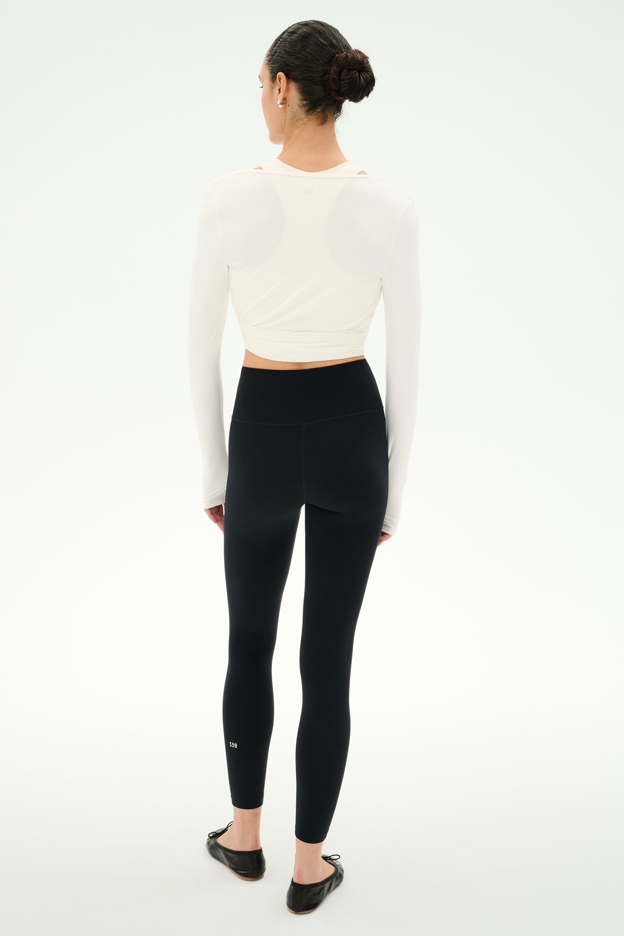 A person stands facing away, wearing the SPLITS59 Bella Airweight Lite Wrap in white, paired with high-waisted black nylon-spandex leggings and black ballet flats, against a plain white background.