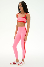 A woman stands in a studio wearing the SPLITS59 Monah Rigor Bra in Strawberry/Lotus Red, paired with high-waisted pink leggings featuring a red stripe and white sneakers with red accents.