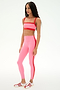 A woman stands in a studio wearing the SPLITS59 Monah Rigor Bra in Strawberry/Lotus Red, paired with high-waisted pink leggings featuring a red stripe and white sneakers with red accents.