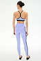 A woman faces away, wearing the SPLITS59 Emerson Airweight Bra in Purple Haze/Black and high-waisted lavender leggings with black side stripes, made from stretch fabric, set against a plain white background.
