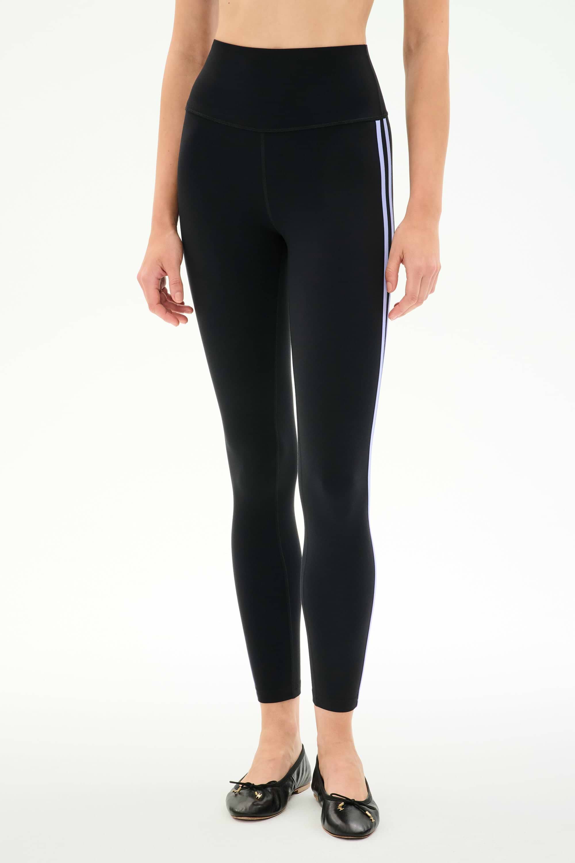 A person wears SPLITS59 Ella High Waist Airweight 7/8 leggings in Black/Purple Haze, featuring two side stripes, with black ballet flats, standing against a plain white background.