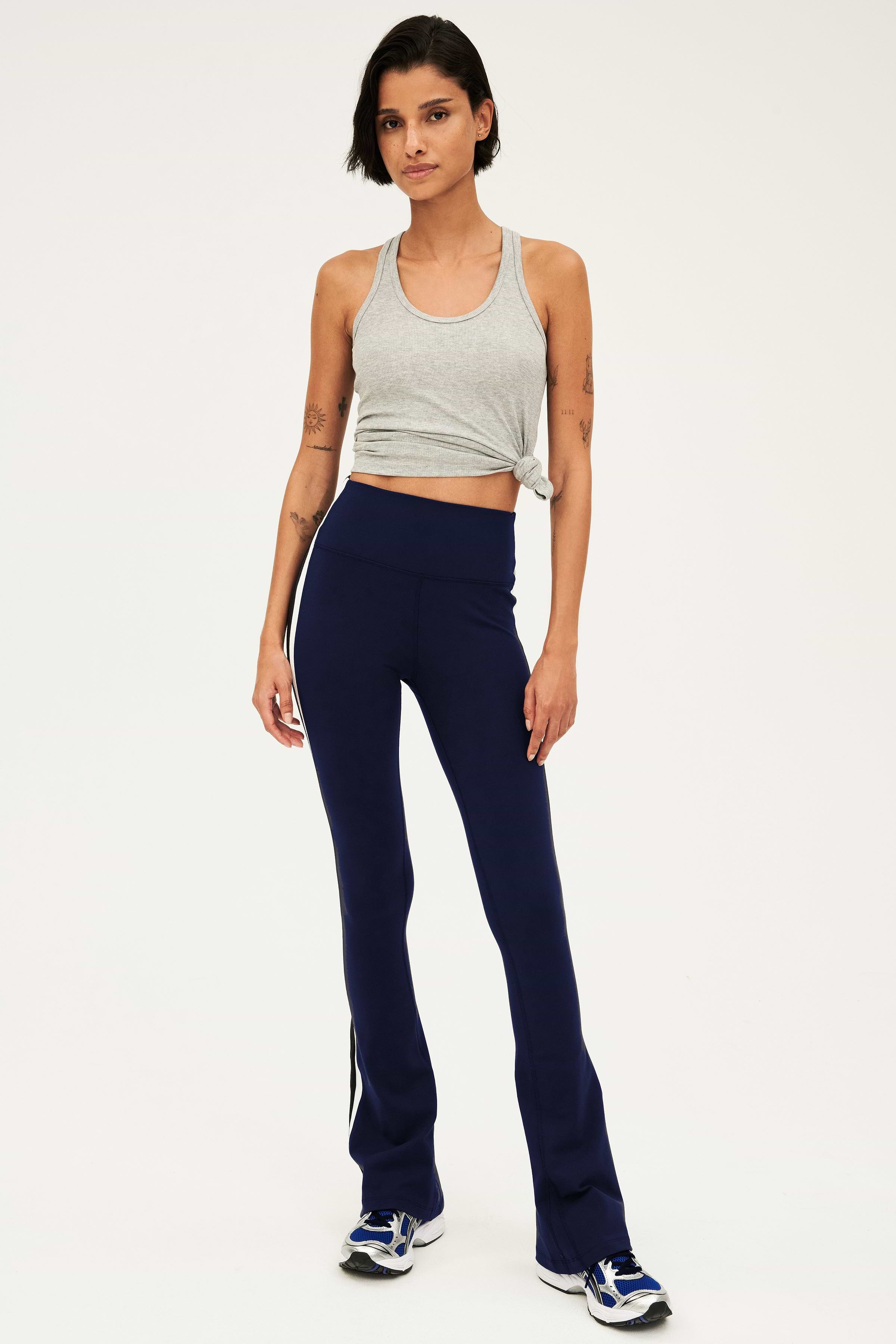 Full front view of woman with dark short hair wearing dark blue high waist below ankle length legging with wide flared bottoms and white and black side stripes on both legs. Also wearing a dark gray racerback tank top with right knot on the bottom. Paired with gray,black and bright blue shoes.