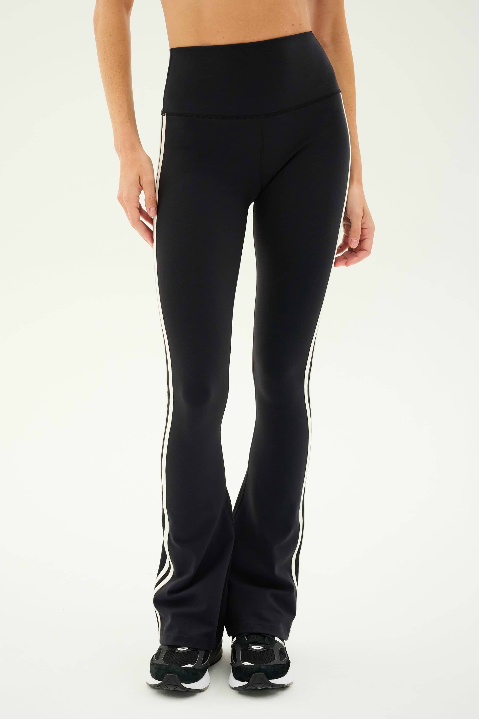 A person wearing the SPLITS59 Raquel High Waist Flared Legging in Black/White paired with black sneakers stands against a plain background.