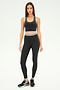 A woman is standing against a plain background, wearing the Bailey Active Rib Bralette in Heather Graphite from SPLITS59 paired with matching leggings, complemented by black sneakers. Her long brown hair cascades down her back, framing a neutral expression that hints at focus—perhaps ready for a Pilates session.
