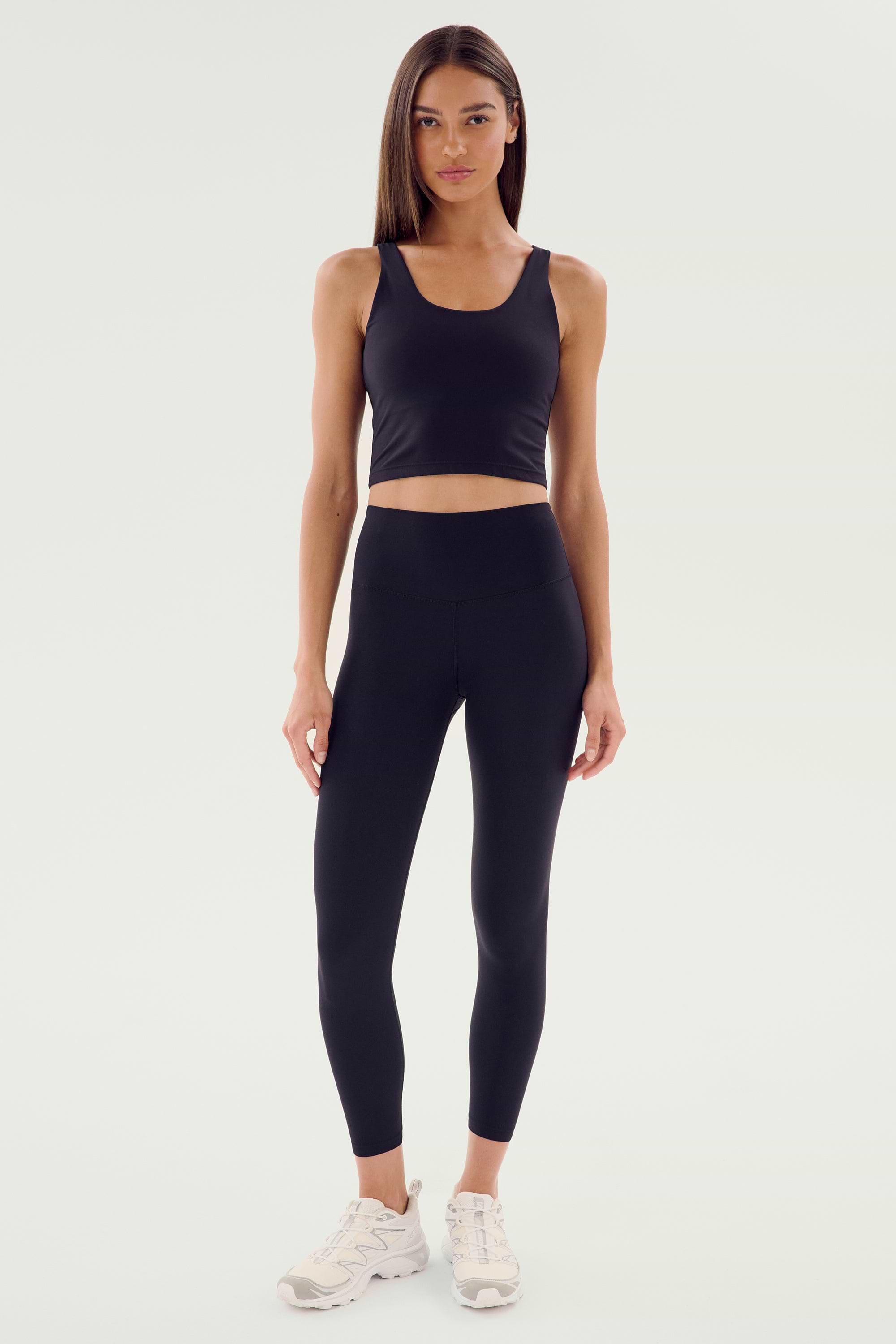 A woman is standing in a black tank top, wearing Sprint High Waist Rigor 7/8 leggings from SPLITS59 with white sneakers, posing against a plain, light-colored background.