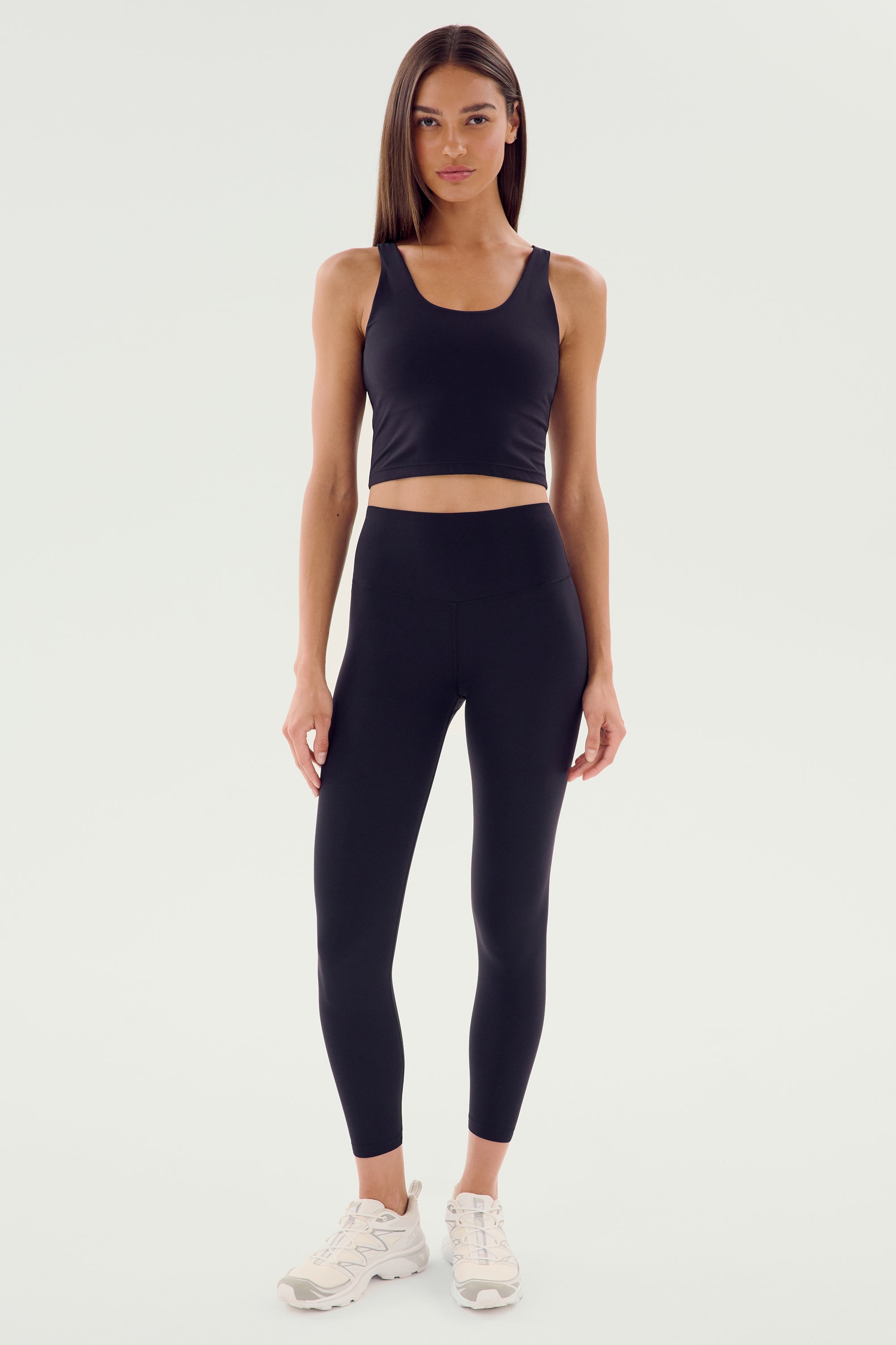 A woman is standing in a black tank top, wearing Sprint High Waist Rigor 7/8 leggings from SPLITS59 with white sneakers, posing against a plain, light-colored background.