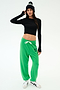 Woman wearing a black long-sleeve crop top, green sweatpants with a drawstring and "59" print, white sneakers, and a black beanie poses against a plain background.