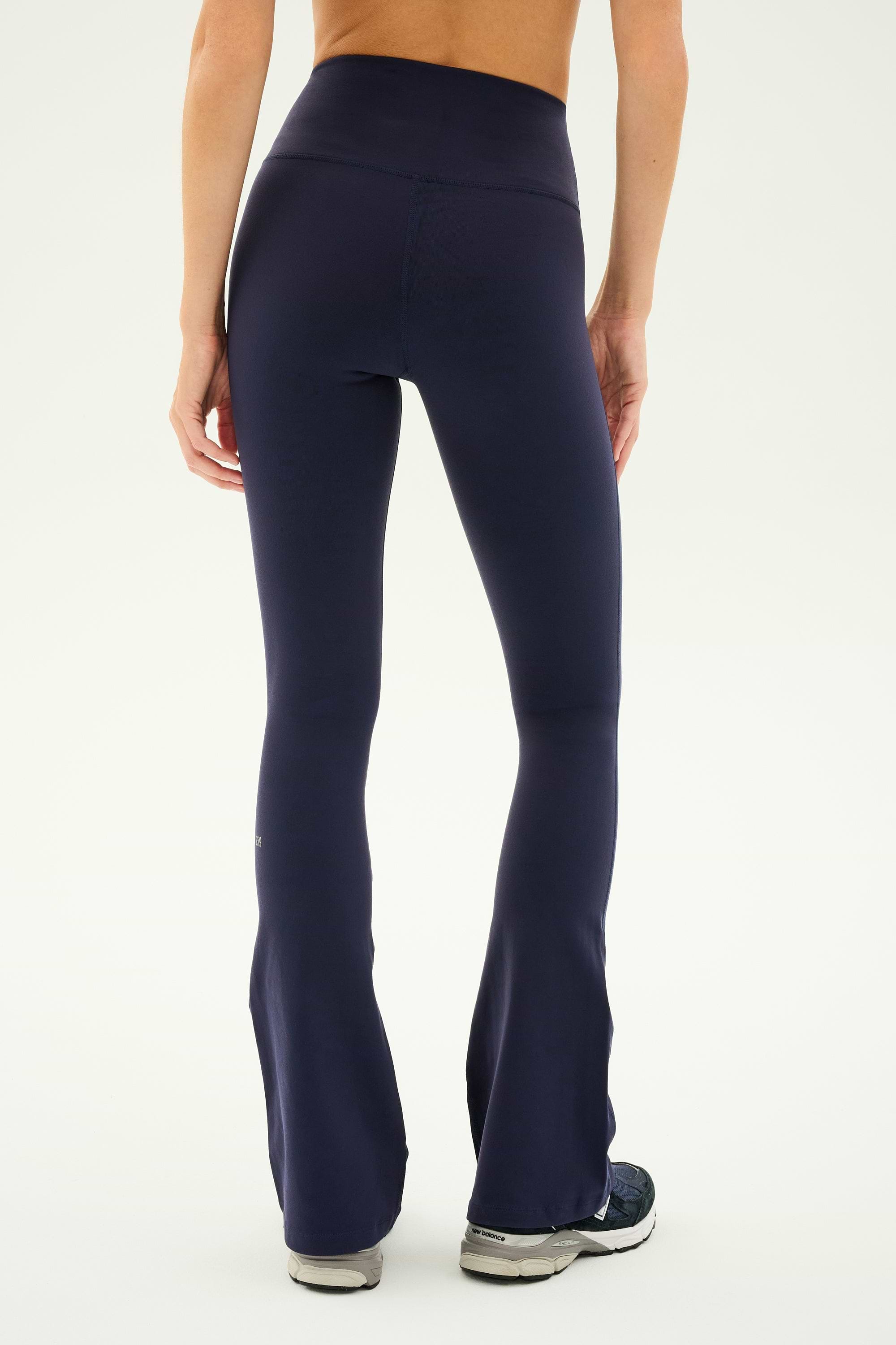 A person displays the back view of their outfit, wearing SPLITS59's Raquel High Waist Flared Legging in Indigo and gray athletic shoes against a plain off-white background.