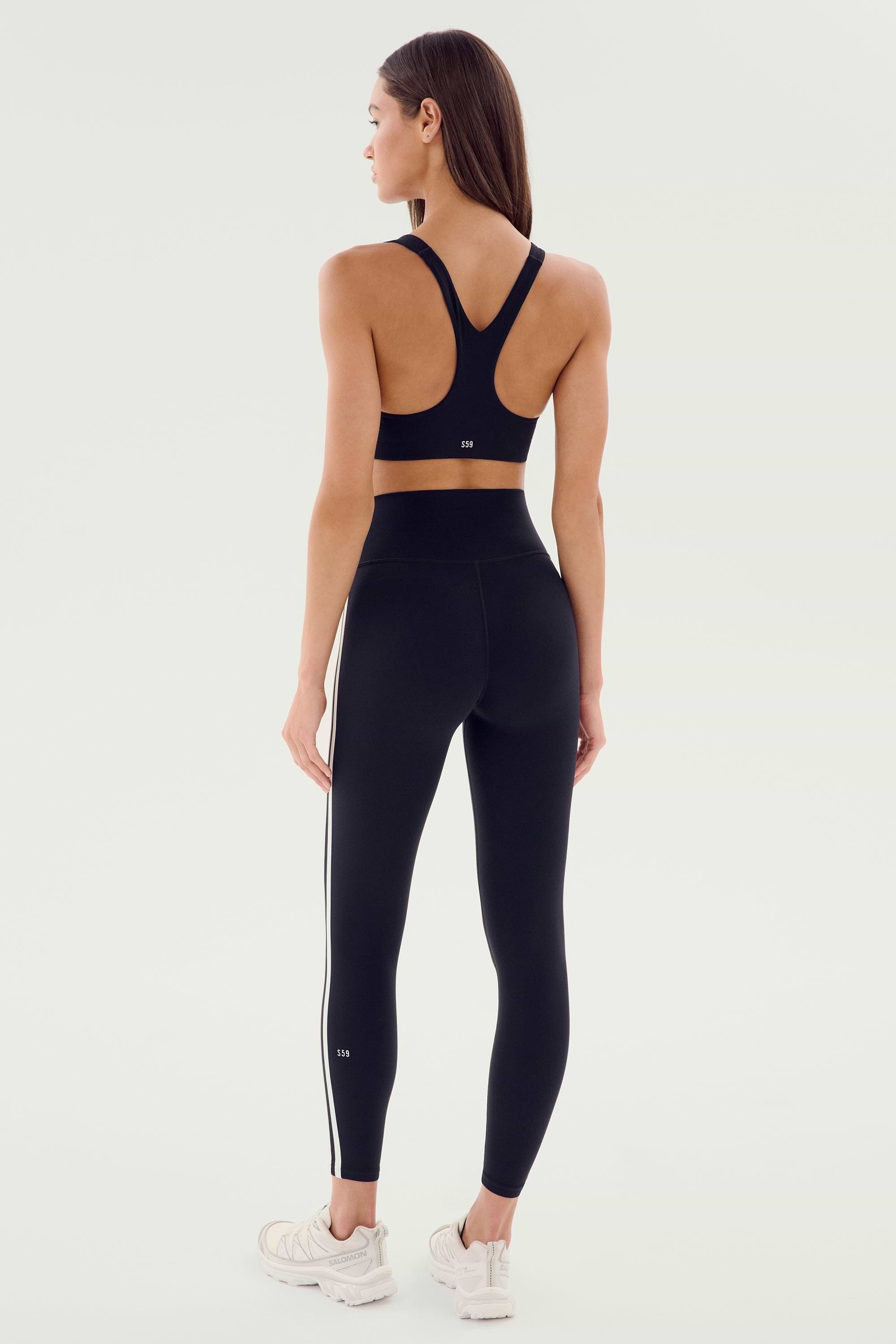 A person stands facing away, wearing the SPLITS59 Ella Airweight Bra in Black/White, high-waisted black leggings with white stripes, and white athletic shoes, ready for a hot yoga session.