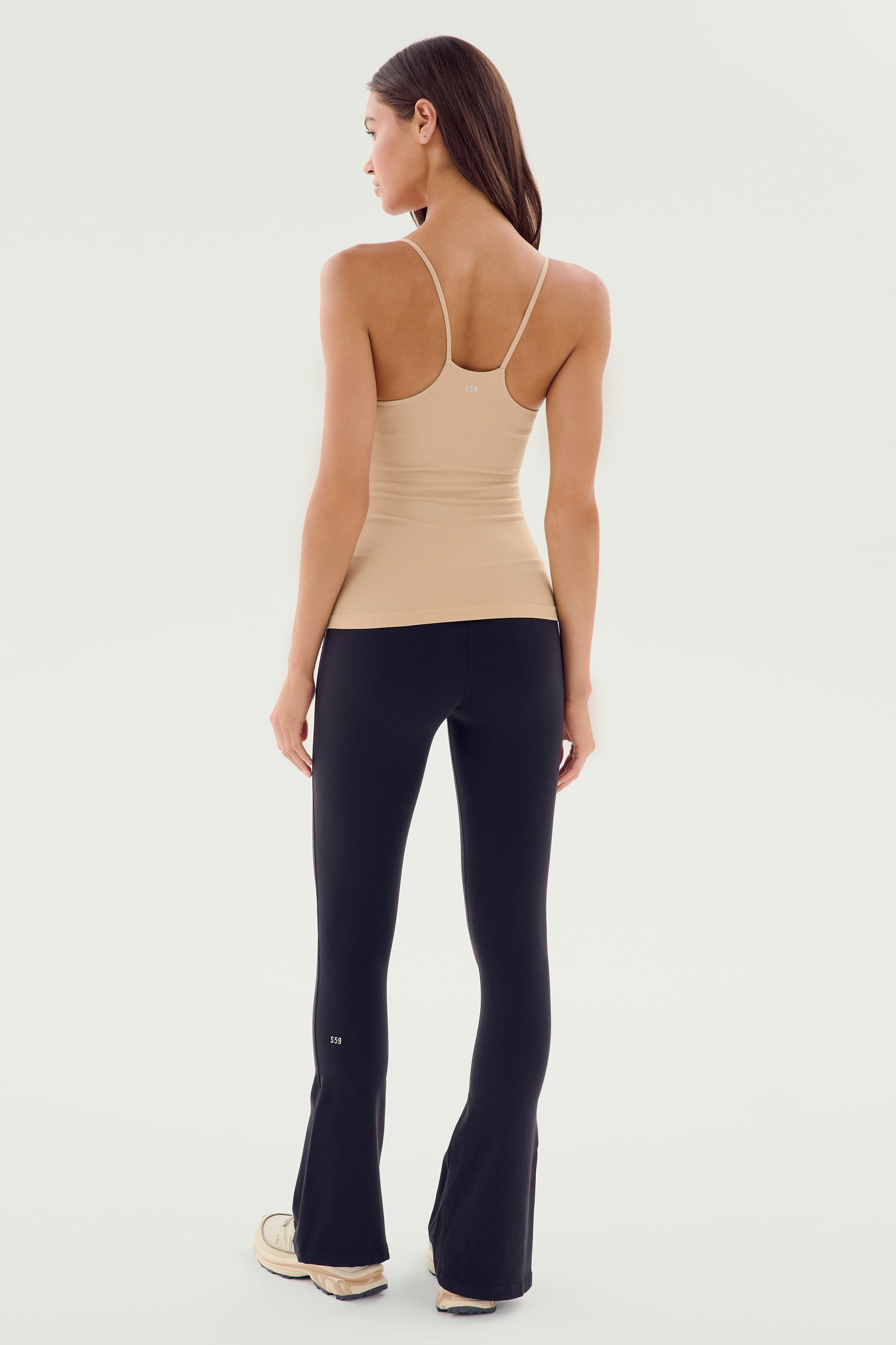 A woman is standing, facing away, wearing the SPLITS59 Loren Seamless Waist Length Tank in Nude, black pants, and white sneakers.
