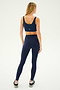 A woman with long blonde hair stands facing away, wearing a SPLITS59 Sprint Rigor Bra in Indigo, high-waisted leggings, and black sneakers against a plain light background. Her outfit exemplifies multi-sport performance, ideal for diverse gym workouts.