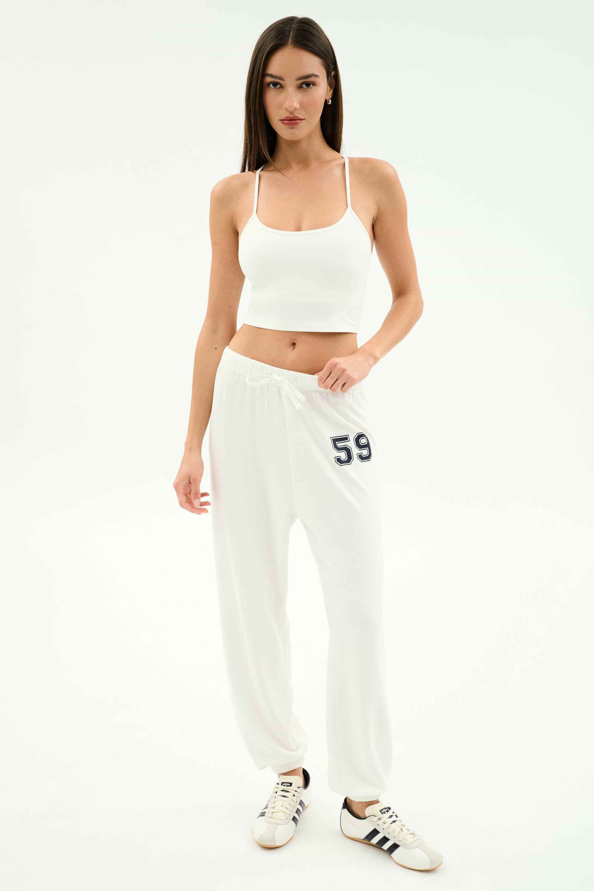 A woman wears the SPLITS59 Airweight Crop - White with white sweatpants featuring 59 and white Adidas sneakers, standing against a plain white background.
