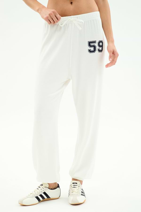 A person wears SPLITS59's Andie Oversized Fleece Varsity 59 Sweatpant in White/59, featuring the number 59 on the left hip, paired with white sneakers with black stripes—ideal for cool weather workouts—against a plain white background.