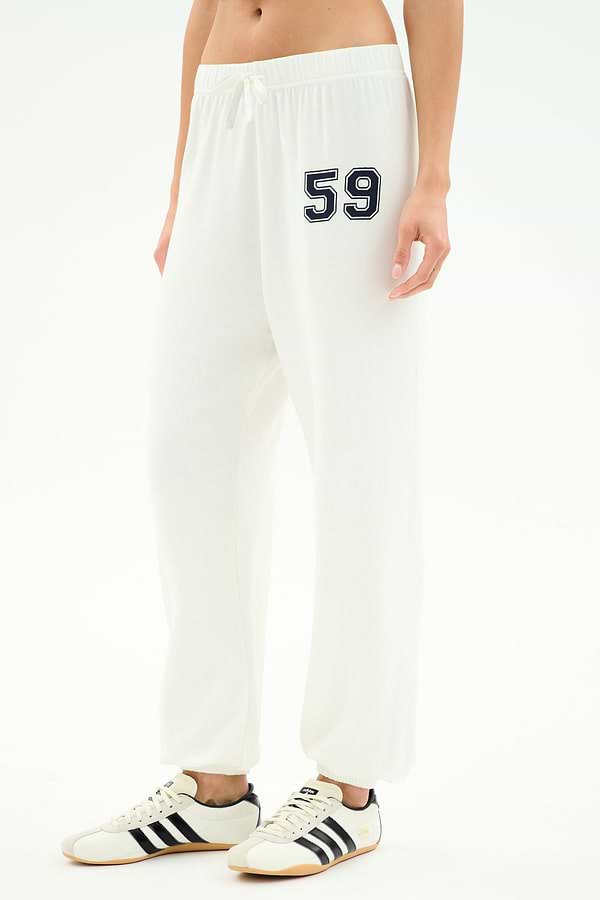 A person in SPLITS59 Andie Oversized Fleece Varsity 59 Sweatpant—white with bold "59"—paired with white sneakers with black stripes, perfect for cool weather workouts. Made in Los Angeles.