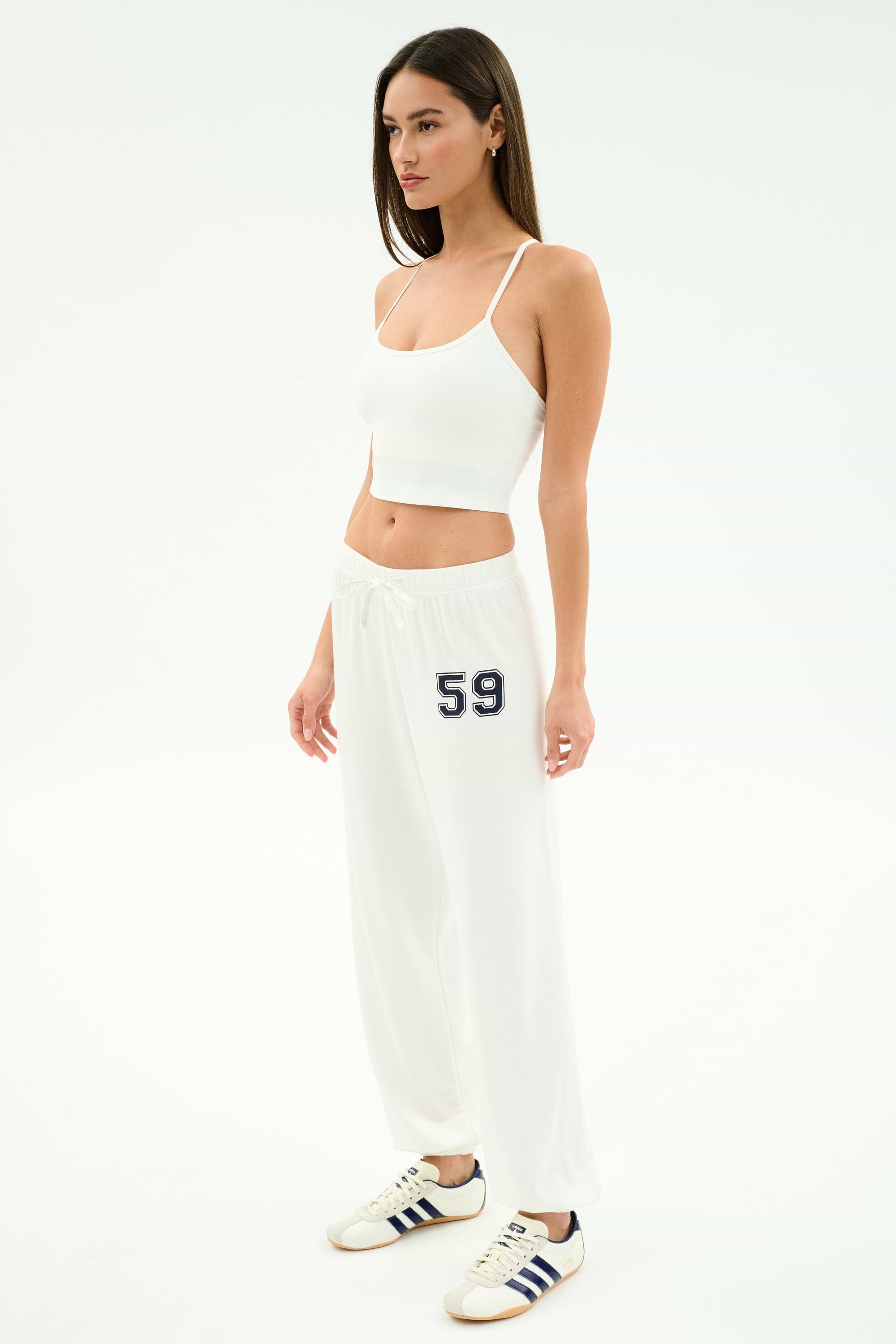 A woman poses in a white studio wearing the SPLITS59 Airweight Crop - White, matching drawstring pants with "59" on the left leg, and white sneakers with navy stripes—ideal for hot yoga or a casual outing.