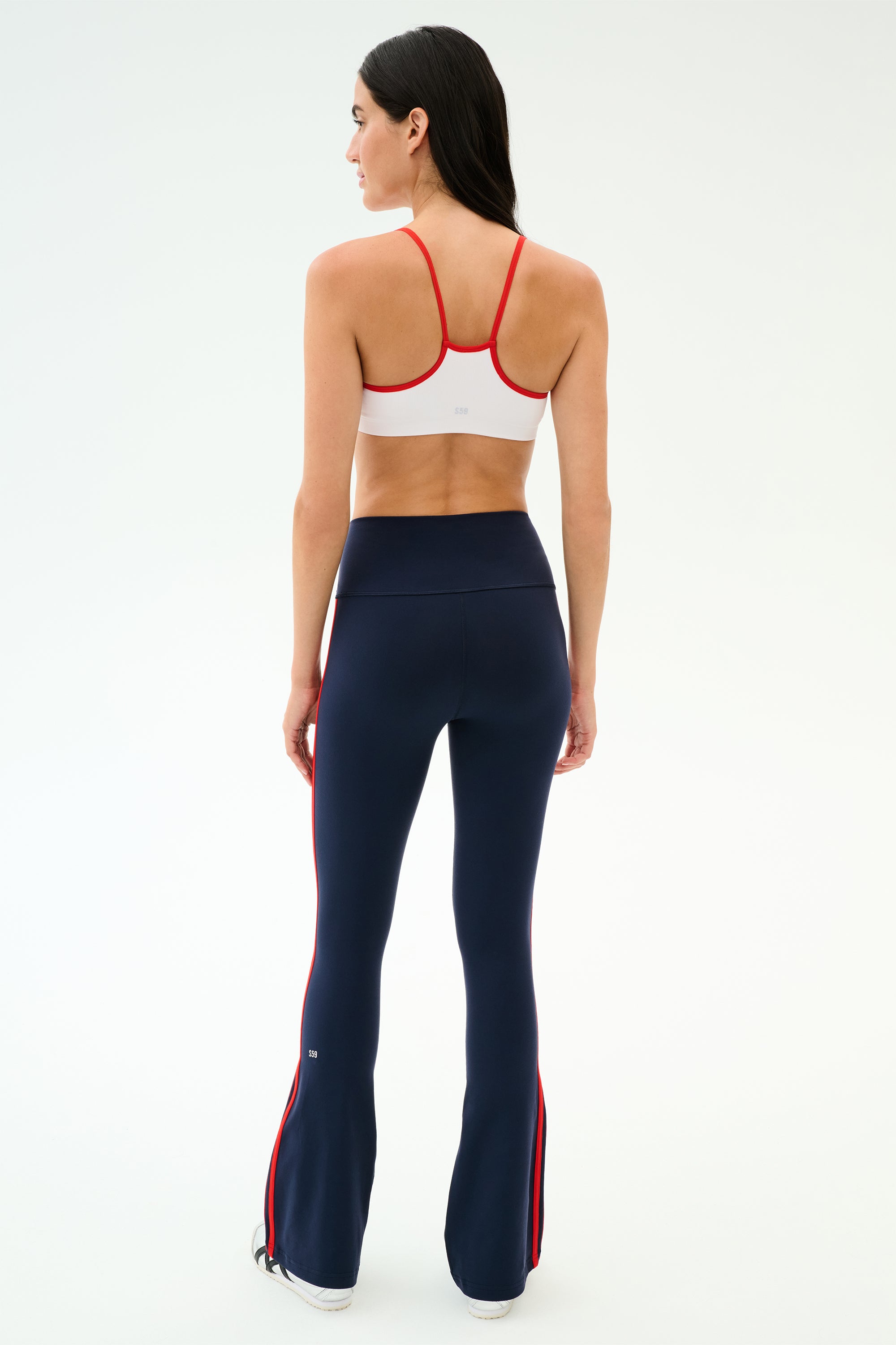 A woman faces away wearing the SPLITS59 Loren Seamless Bra in White/Lotus Red, paired with high-waisted navy flared leggings featuring red side stripes—an ideal yoga or gym outfit on a plain light background.