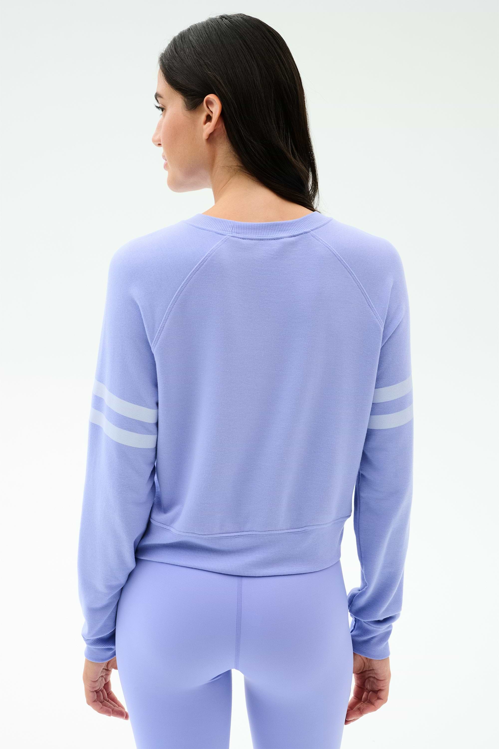 A woman with long dark hair wears the SPLITS59 Brooks Stripe Fleece Sweatshirt in Purple Haze/White, featuring two white sleeve stripes, made in Los Angeles, standing with her back to the camera.