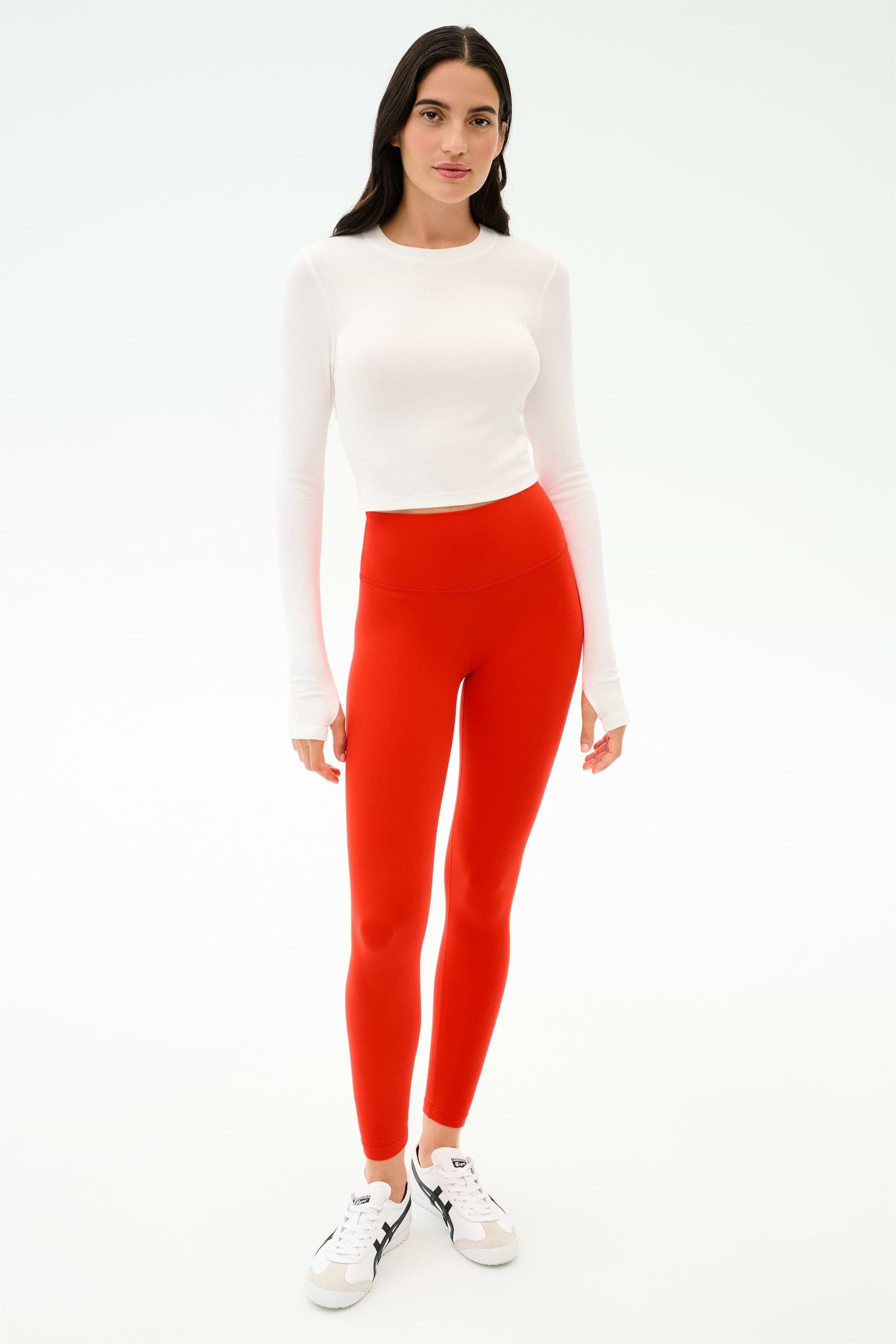 A person stands facing forward in a white long-sleeve top, white sneakers, and SPLITS59 Sprint High Waist Rigor 7/8 leggings in Lotus Red, against a plain white background.