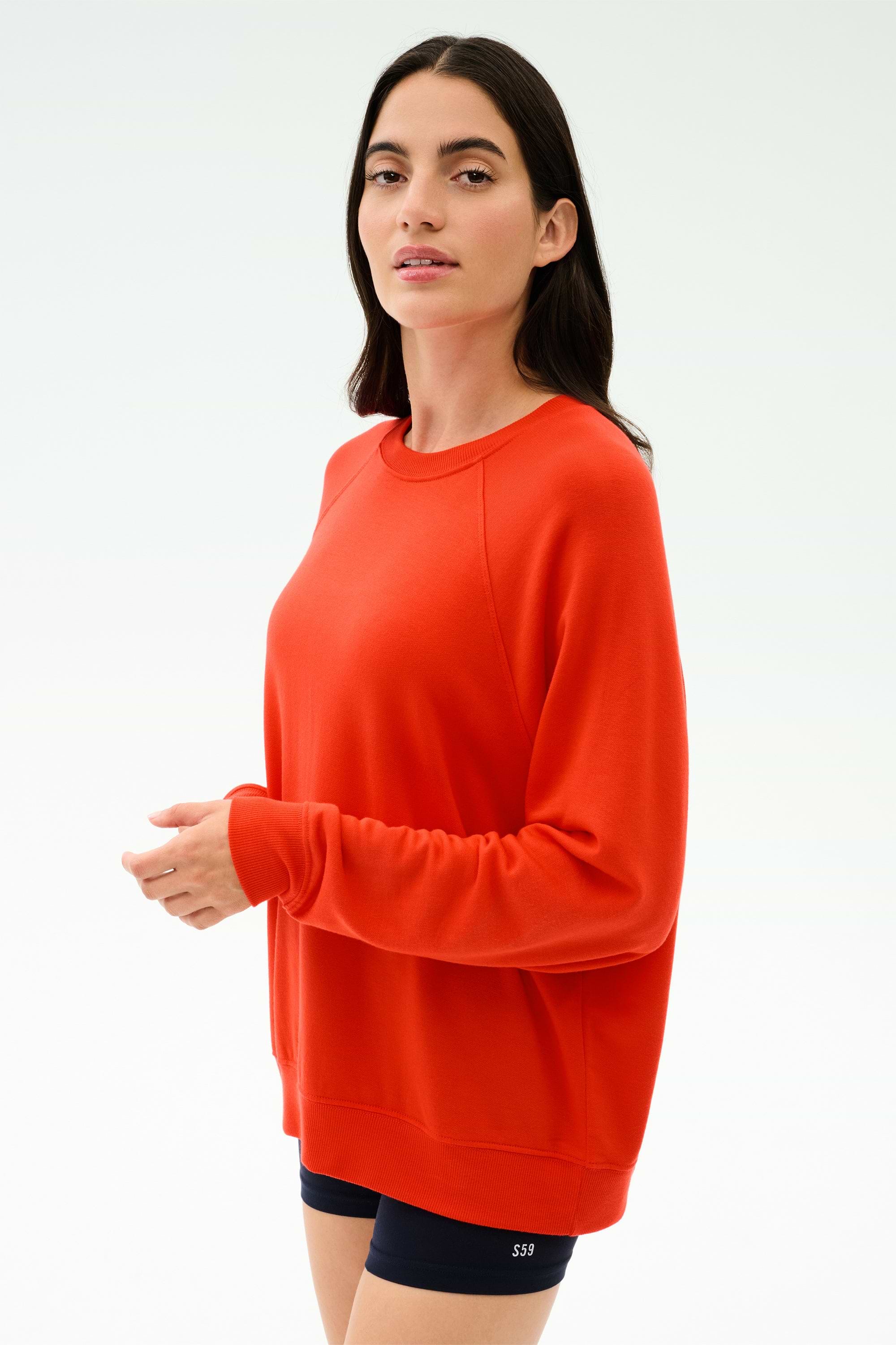 A woman in the SPLITS59 Andie Oversized Fleece Sweatshirt in Lotus Red and black shorts, made in Los Angeles, stands against a light background, facing the camera.