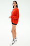 A woman stands against a plain background wearing the SPLITS59 Andie Oversized Fleece Sweatshirt in Lotus Red, paired with black shorts, white socks, and white sneakers.