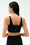 A woman with long dark hair, seen from behind, wears the SPLITS59 Sprint Rigor Bra in black and leggings, ready for her gym workout against a plain light background.
