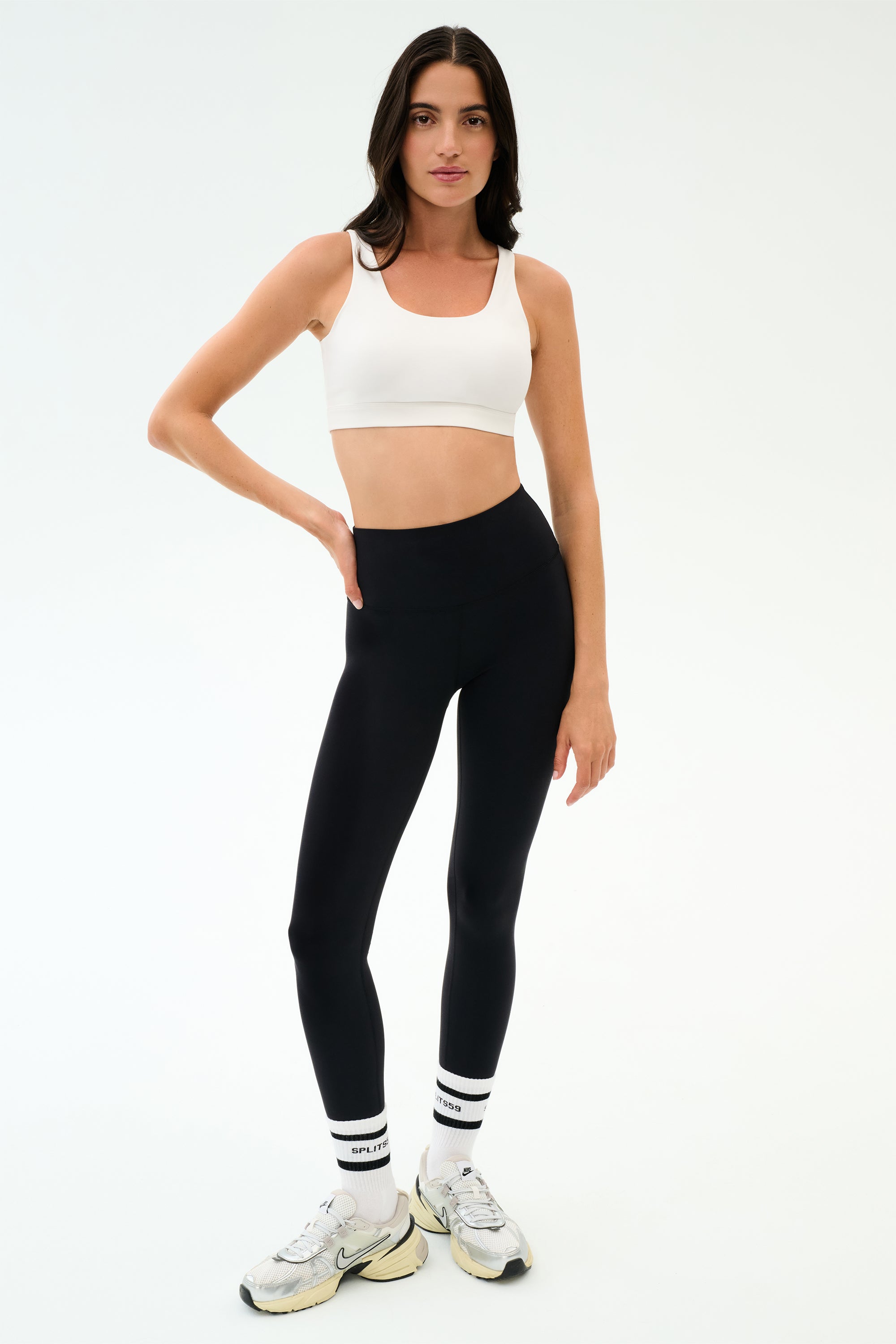A woman stands against a plain background wearing the SPLITS59 Sprint Rigor Bra in White, black leggings, white socks, and white sneakers—perfect for gym sessions or multisport performance.