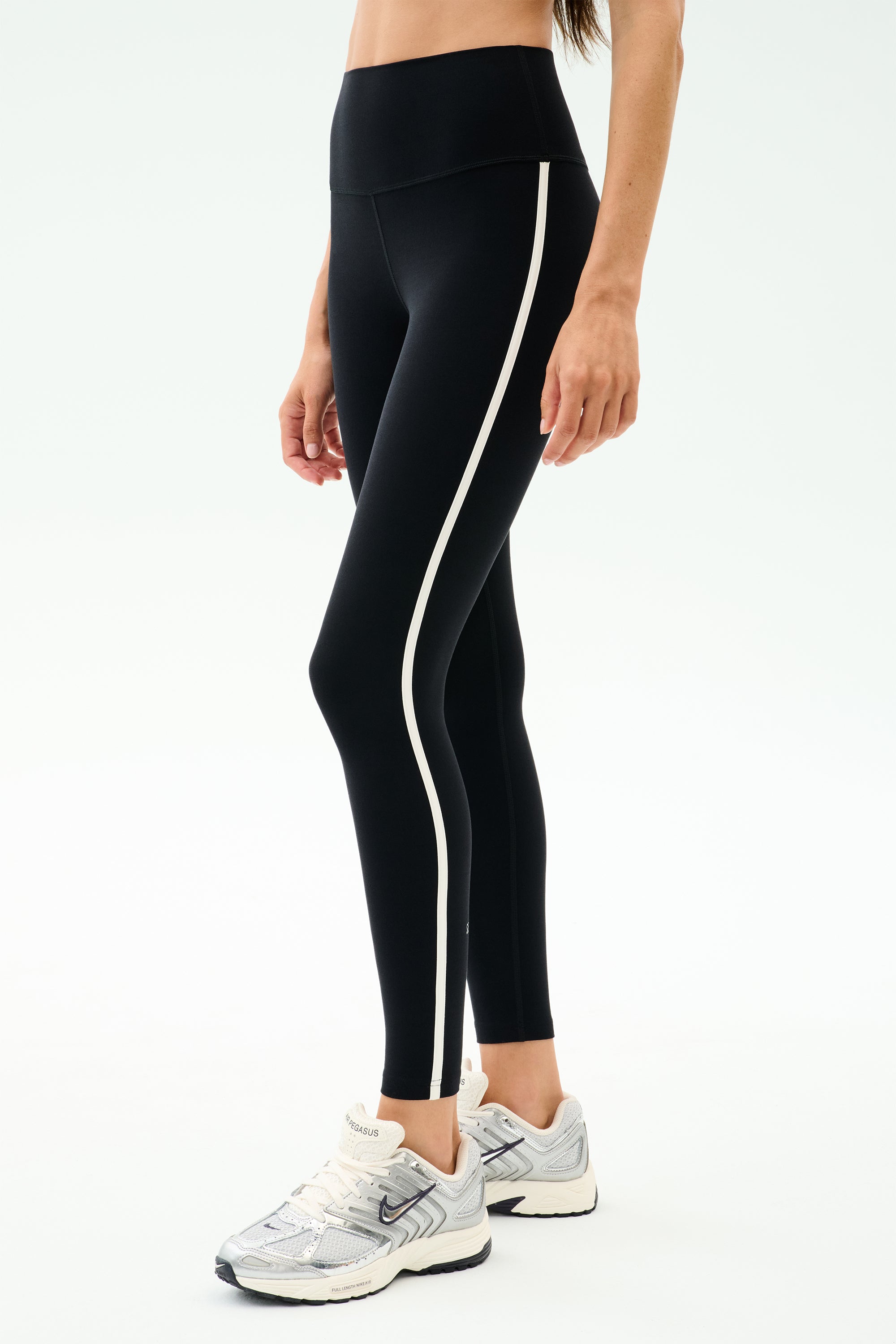 Wearing SPLITS59 Amber Airweight High Waist 7/8 leggings in Black/White with side stripes and white sneakers—ideal for hot yoga or casual wear against a plain backdrop.