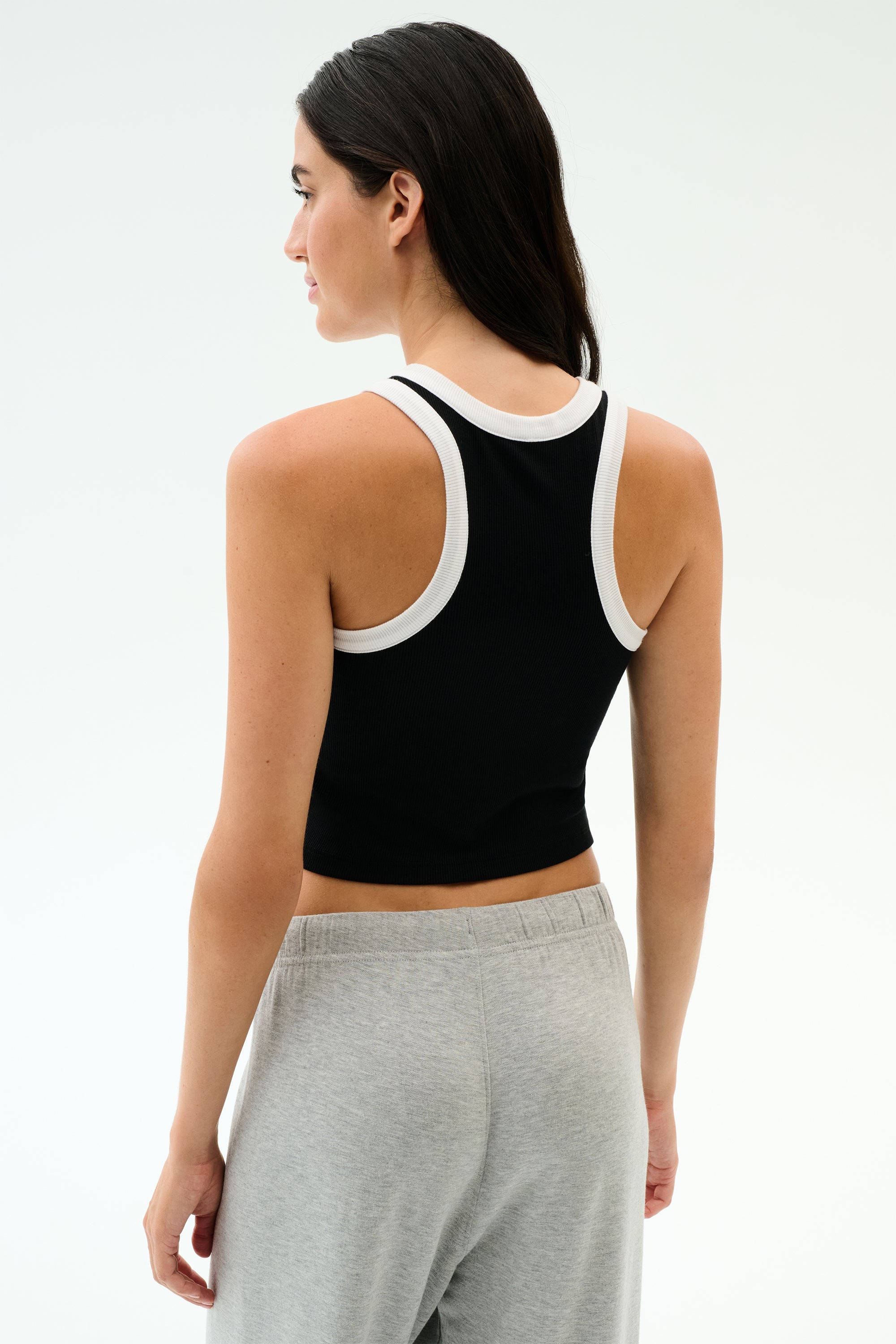 A woman with long dark hair stands with her back to the camera, wearing the SPLITS59 Kiki Rib Crop Tank in Black/White and gray sweatpants.