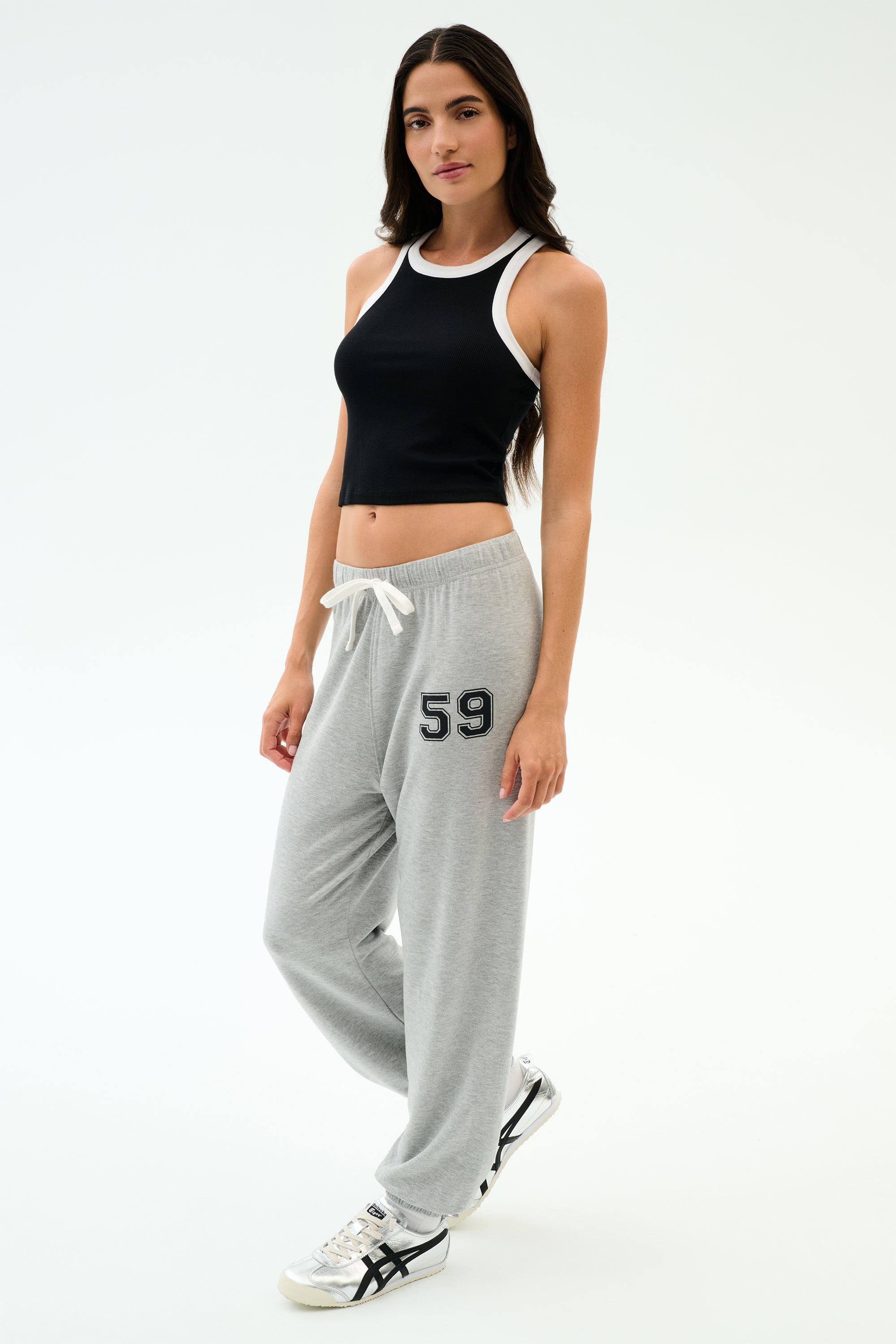 A woman wears the SPLITS59 Kiki Rib Crop Tank in Black/White with light gray sweatpants featuring "59" on the leg and white athletic shoes, set against a plain white background—ideal for gym or casual wear.
