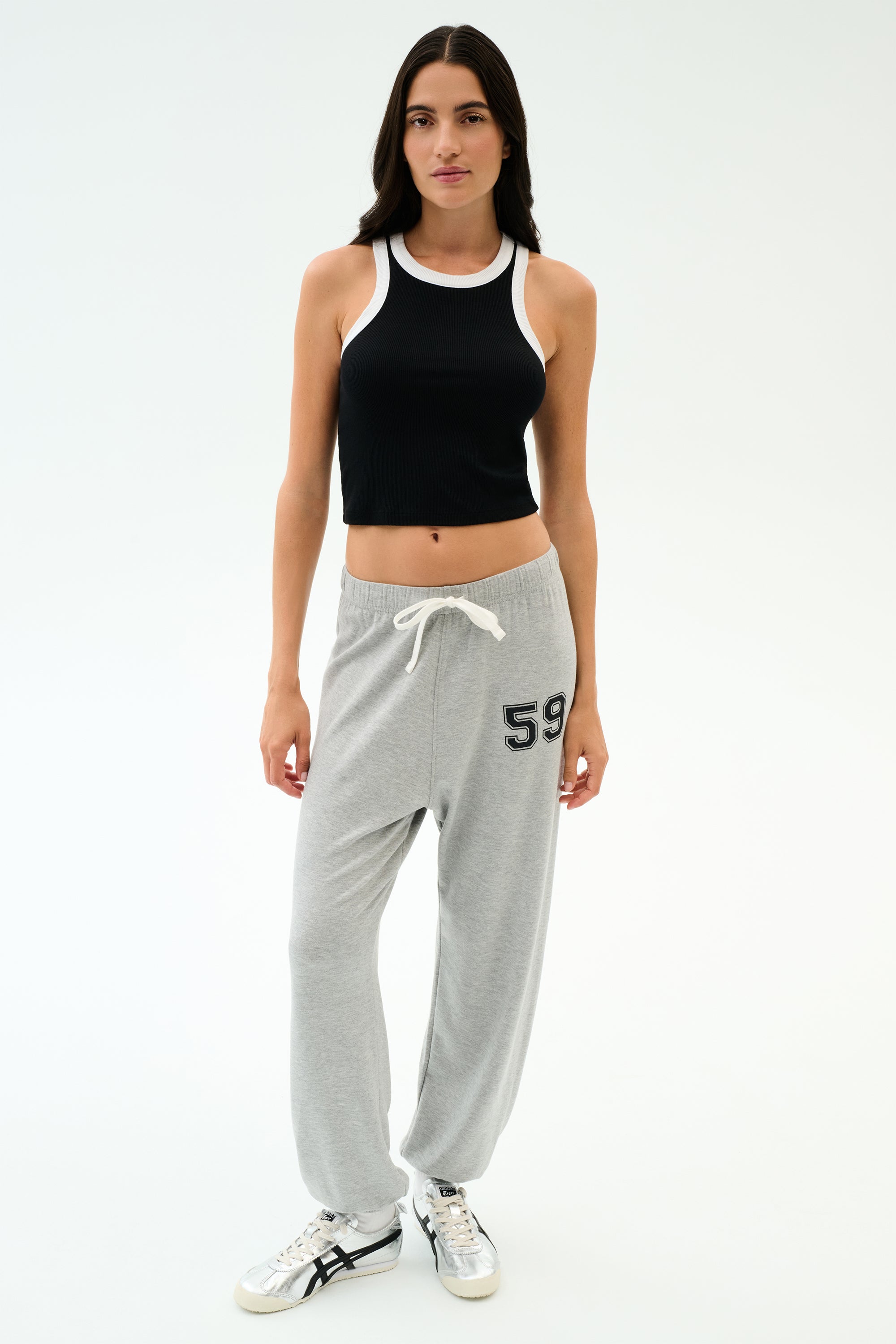 A woman in the SPLITS59 Kiki Rib Crop Tank - Black/White, gray sweatpants with "59" on the left leg, and white sneakers poses against a plain white background—an ideal sporty gym look.