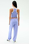 A woman stands facing away, wearing SPLITS59’s Andie Oversized Fleece Sweatpant in Purple Haze with a light purple tank top and white sneakers against a plain backdrop—perfect for warm weather workouts.