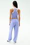 A woman stands facing away, wearing SPLITS59’s Andie Oversized Fleece Sweatpant in Purple Haze with a light purple tank top and white sneakers against a plain backdrop—perfect for warm weather workouts.