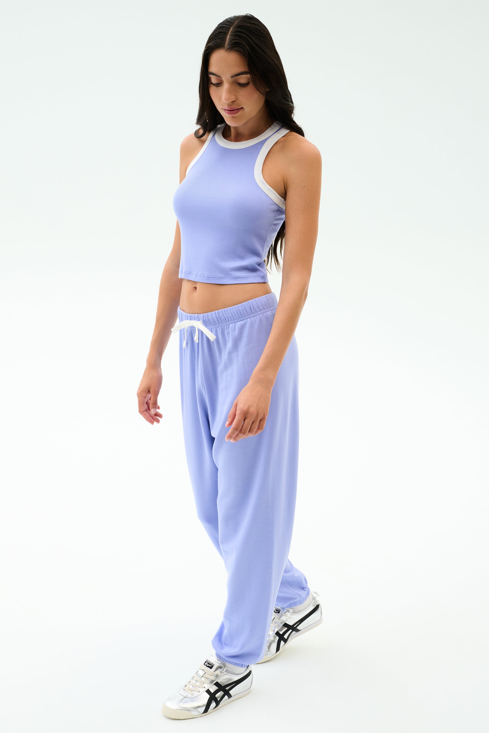 A woman stands against a white background wearing SPLITS59’s Andie Oversized Fleece Sweatpant in Purple Haze, paired with a light purple crop top and white sneakers—ideal for warm weather workouts.