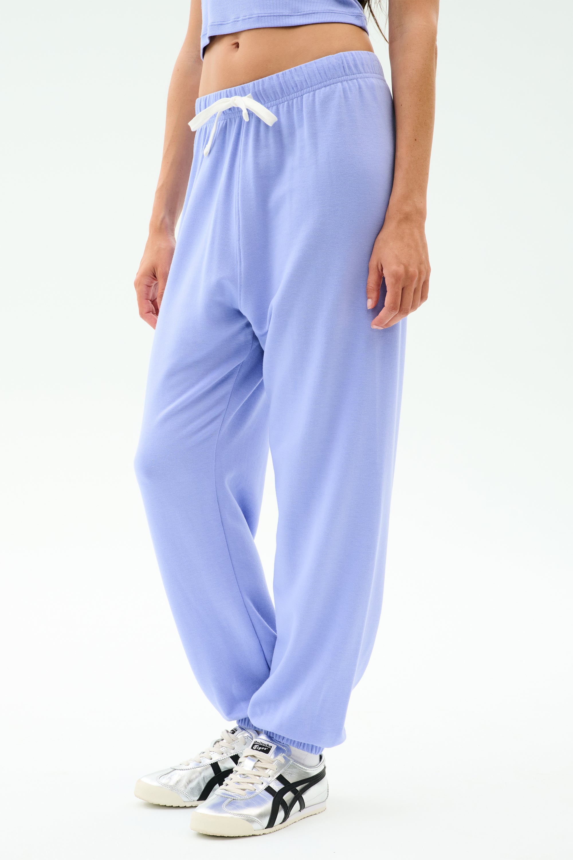 A person wearing SPLITS59 Andie Oversized Fleece Sweatpant in Purple Haze, a cropped top, and white sneakers stands against a plain white background.