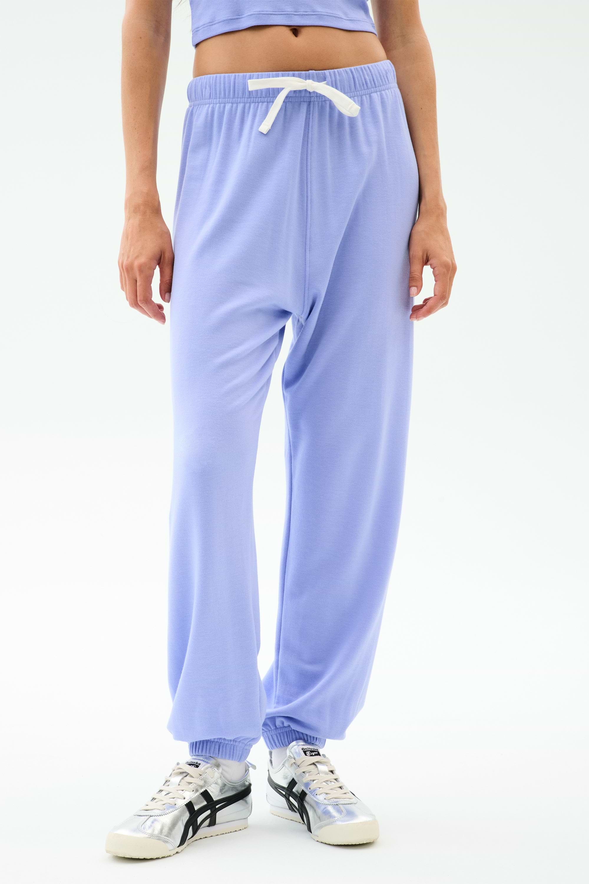 A person models the SPLITS59 Andie Oversized Fleece Sweatpant in Purple Haze—featuring a white drawstring and styled with silver sneakers—against a plain white background, ideal for warm weather workouts.