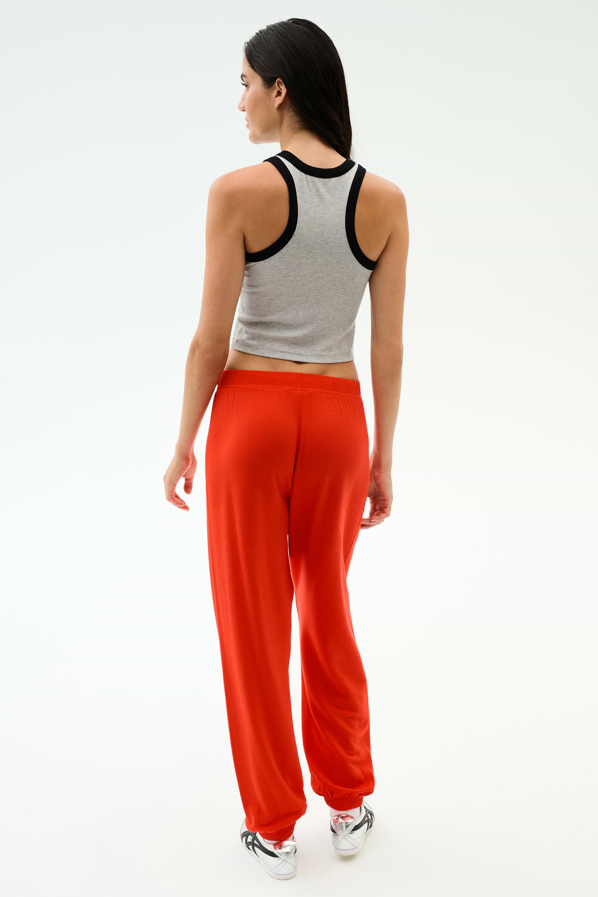 A woman faces away against a white background, wearing a gray tank top, SPLITS59's Andie Oversized Fleece Sweatpant in Lotus Red, and white sneakers—perfect for warm weather workouts.