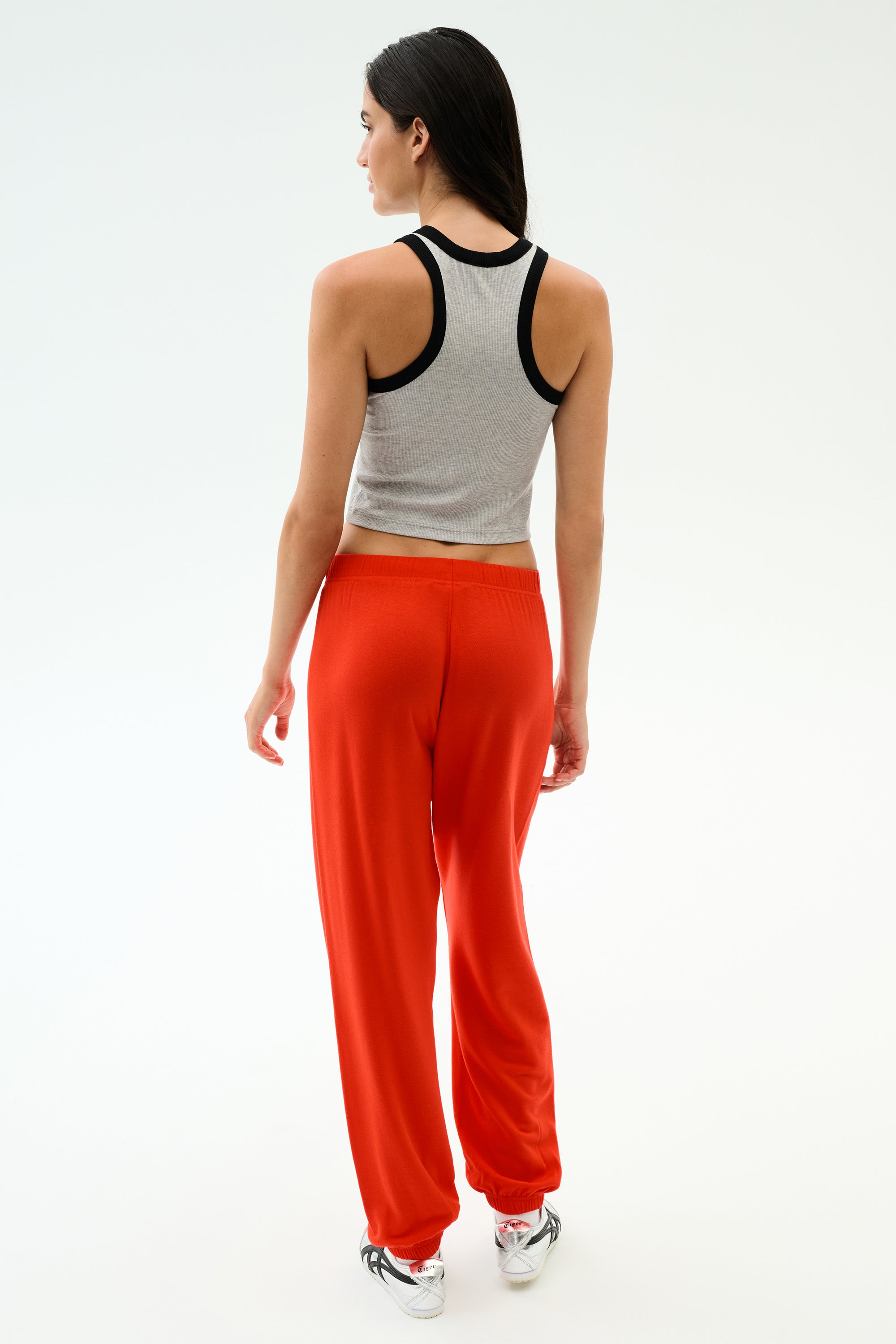 A woman faces away, wearing the SPLITS59 Kiki Rib Crop Tank in Heather Grey/Black, paired with loose red pants and white sneakers, set against a plain light background.
