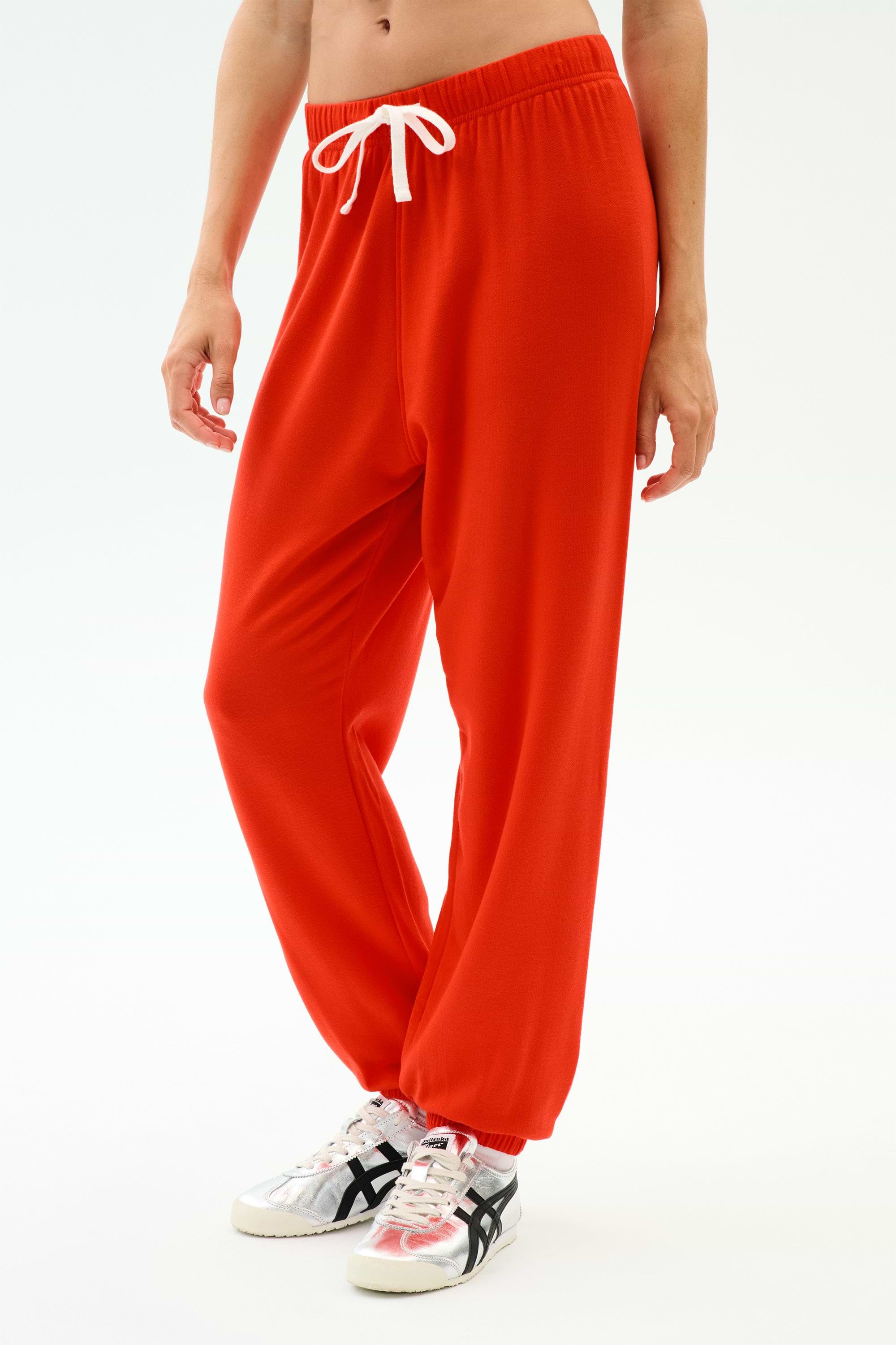 A person wears SPLITS59's Andie Oversized Fleece Sweatpant in Lotus Red with a white drawstring and white sneakers with black stripes, ideal for warm weather workouts, standing against a plain white background.